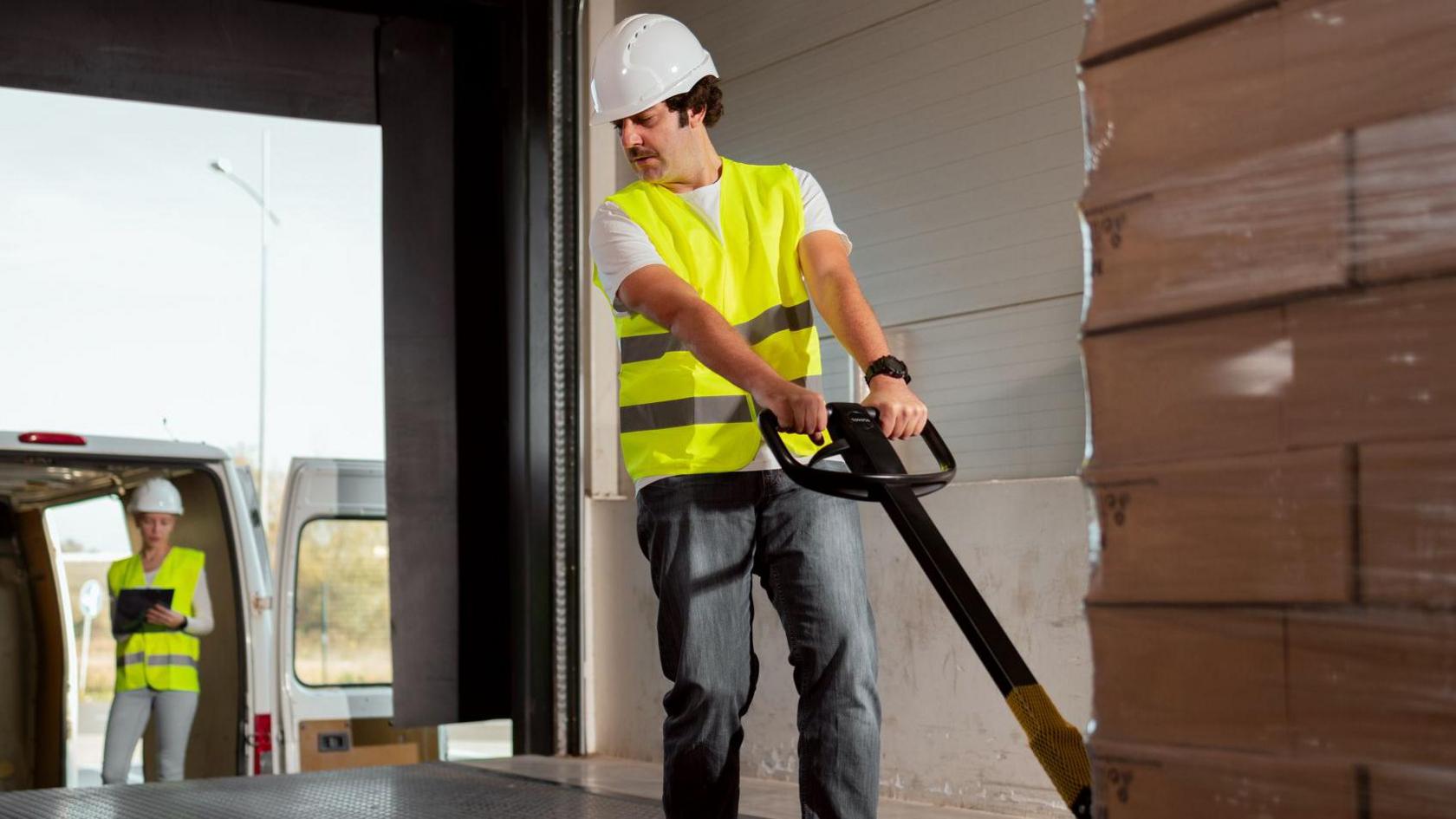 A warehouse worker wearing a hi-vis vest and hard hat pulling a pallet of boxes out of a factory for delivery