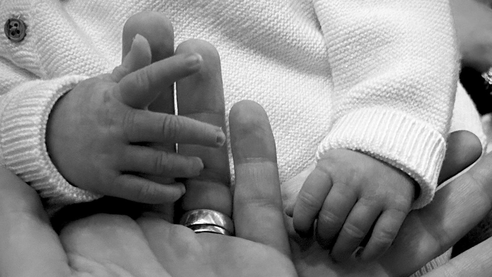 A black and white close up of an adult hand, with a marriage band on its ring finger, holding hands with a newborn. 