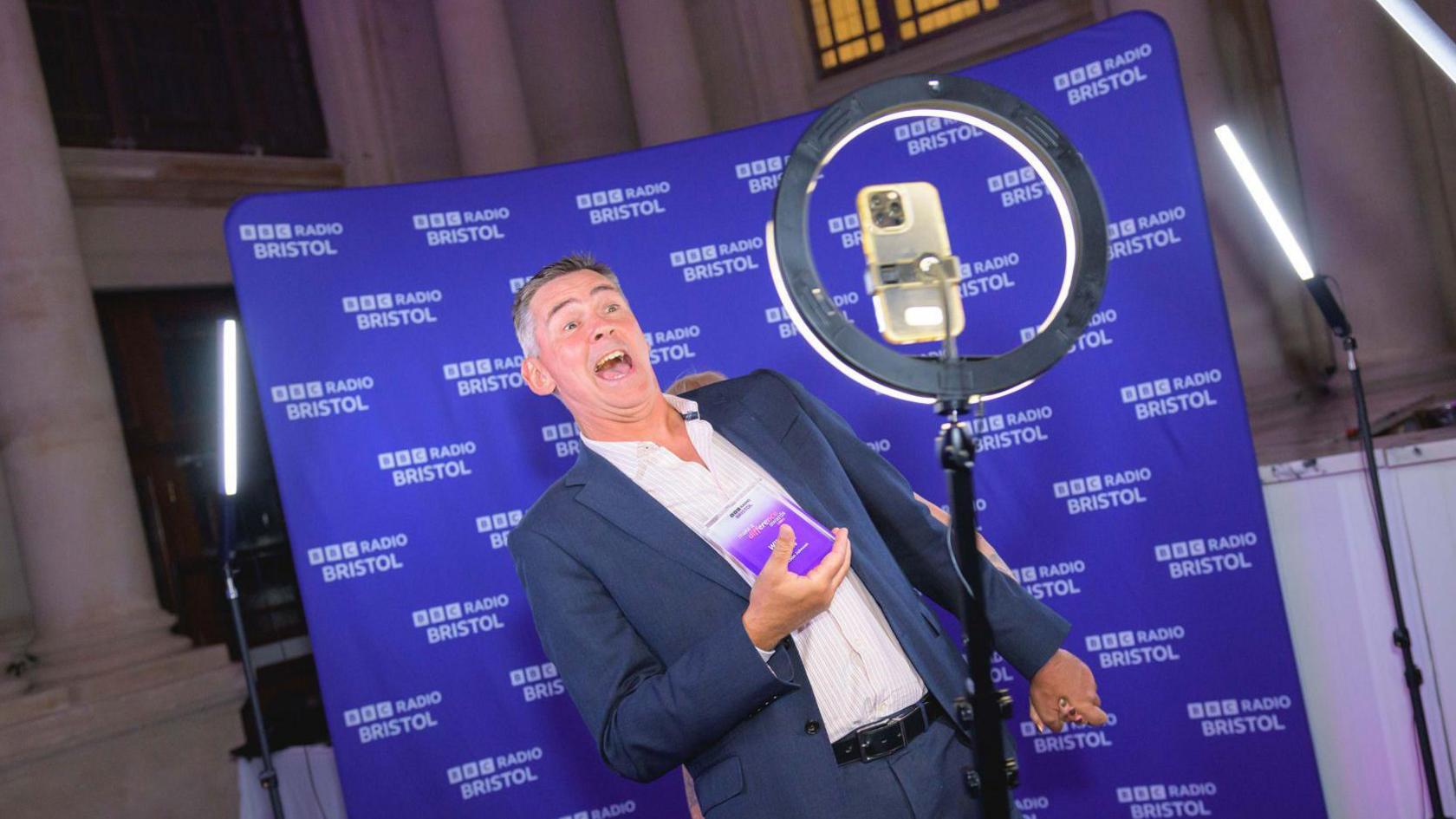 A man leans back while smiling at a phone camera positioned in circular halo light in front of purple background. He is holding his award.