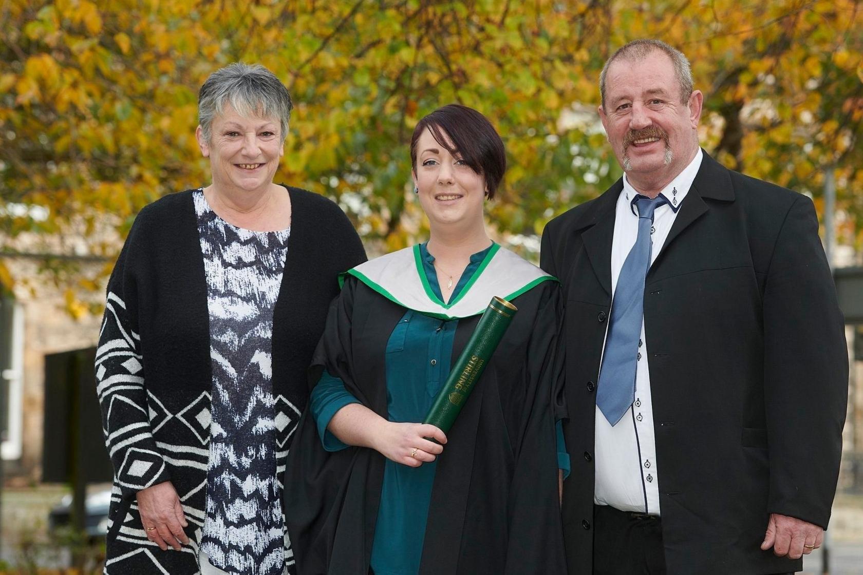 Sheree has short dark hair and wears a green dress and graduation robes and holds a green University of Stirling degree scroll. She is smiling. She is flanked by her parents who are dressed formally and smiling at the camera. Autumn leaves can be seen on the trees in the background.