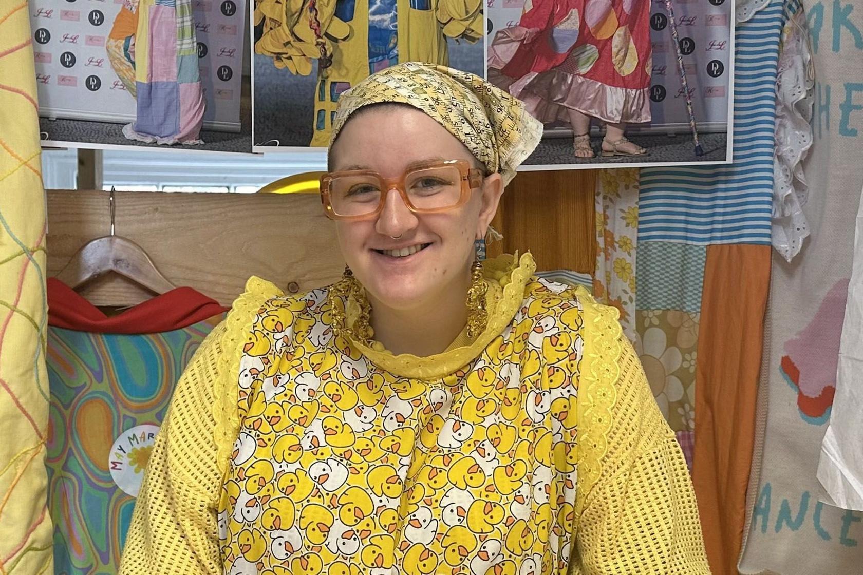 May Gauntlett in her workshop. She is wearing a variety of yellow, patterned clothing, including a headscarf. Fabric and design images are laid on the table and hanging behind her