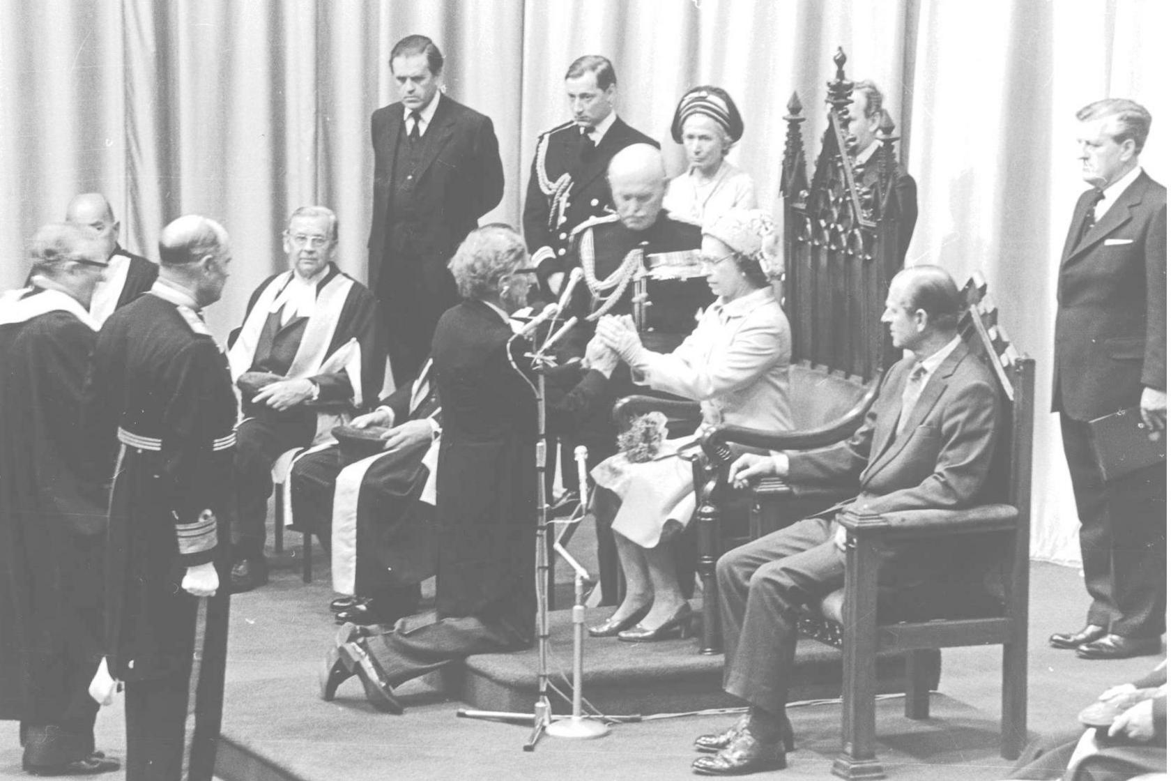 A black and white photo of a young Queen Elizabeth II sitting in a ceremonial wooden chair with several people in suits in military uniforms surrounding her