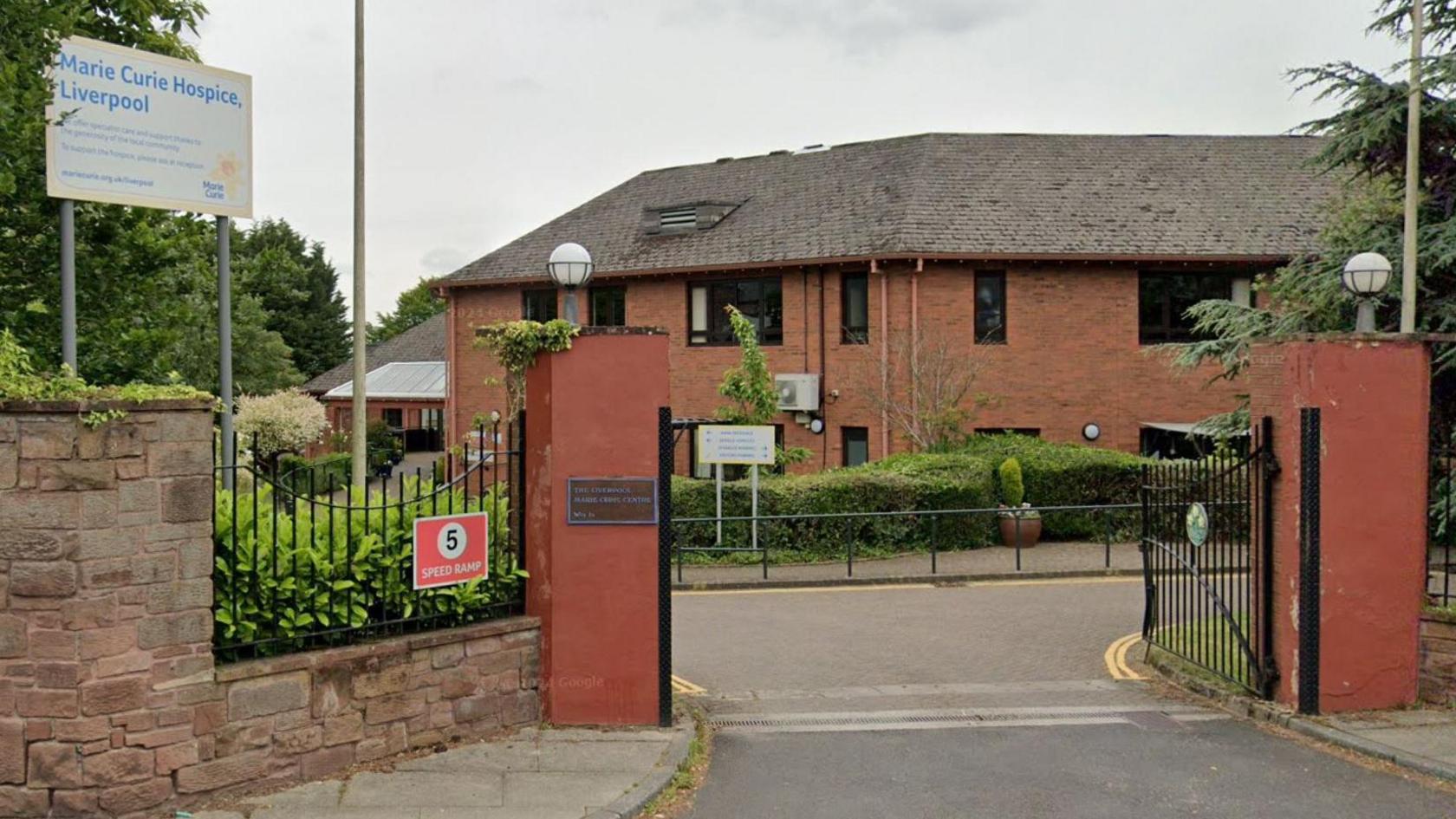 A sandstone wall entrance to the red brick Marie Curie Hospice building surrounded by green trees and bushes