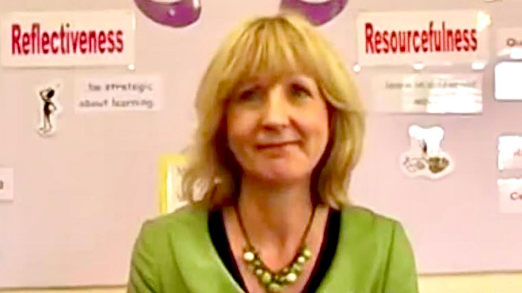 Christine Baker is seen in front of a school noticeboard showing a display about learning strategies. She has blonde hair with a fringe and is wearing a green jacket with a black top and a necklace of green beads.