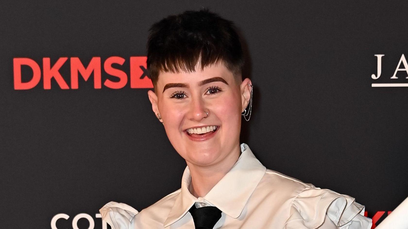 Shell Rowe is standing on a red carpet at an event for DKMS, a blood cancer charity. She is wearing a white shirt and black tie. She is looking at the camera and smiling.