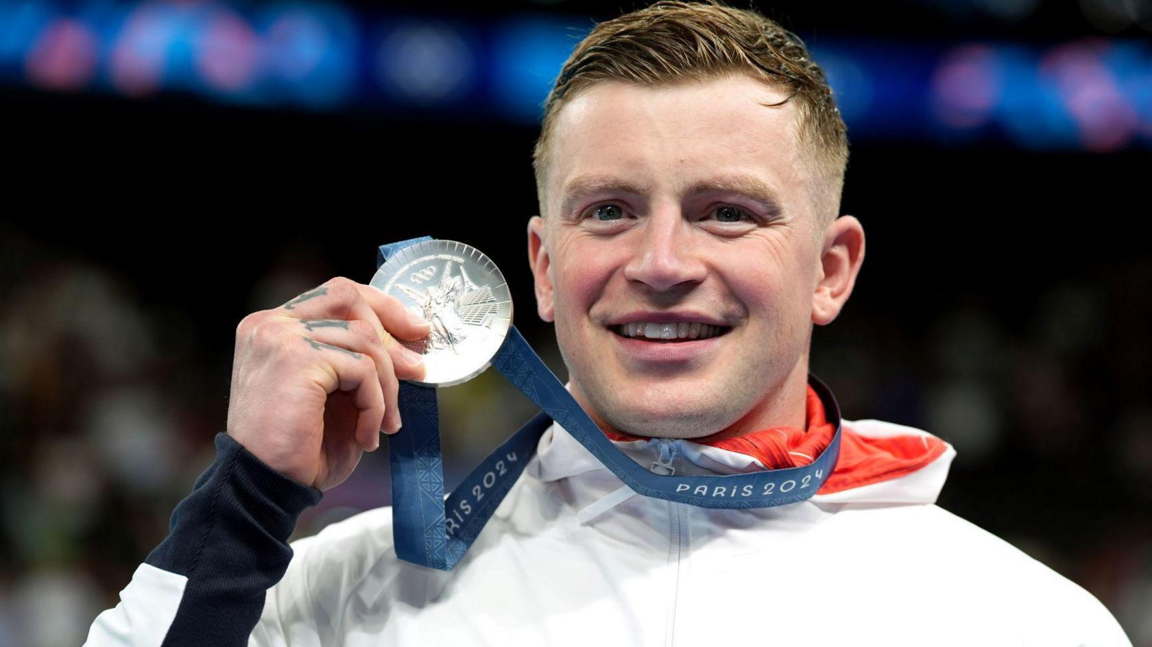 Adam Peaty looking at the camera holding is silver medal in his hand. He has short blond hair and is smiling and wearing a white tracksuit top . 