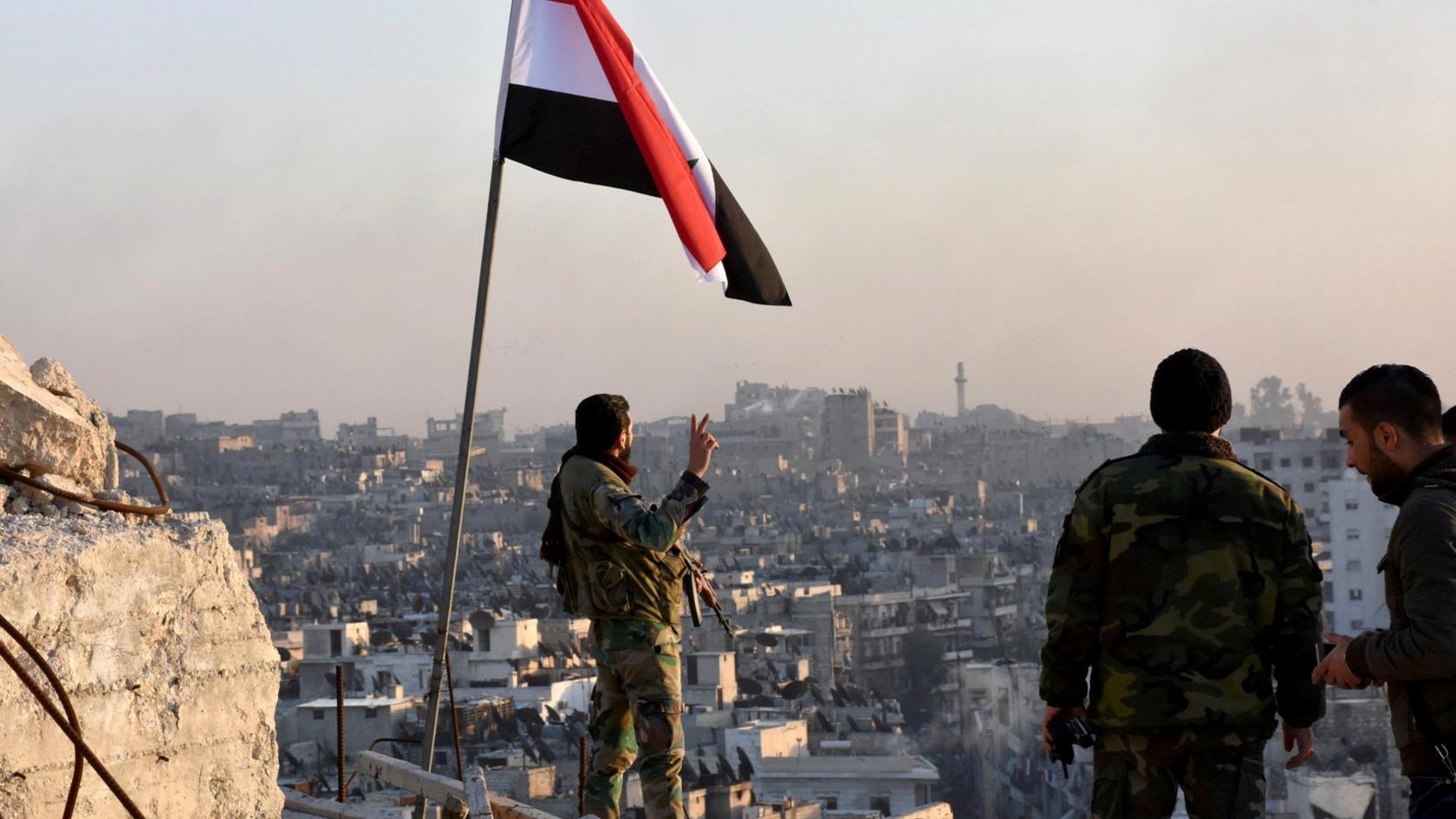 A Syrian government soldier gestures under the Syrian national flag near a general view of eastern Aleppo after they took control of al-Sakhour neigbourhood in Aleppo, Syria in this handout picture provided by SANA Syrian news agency on 28 November 2016