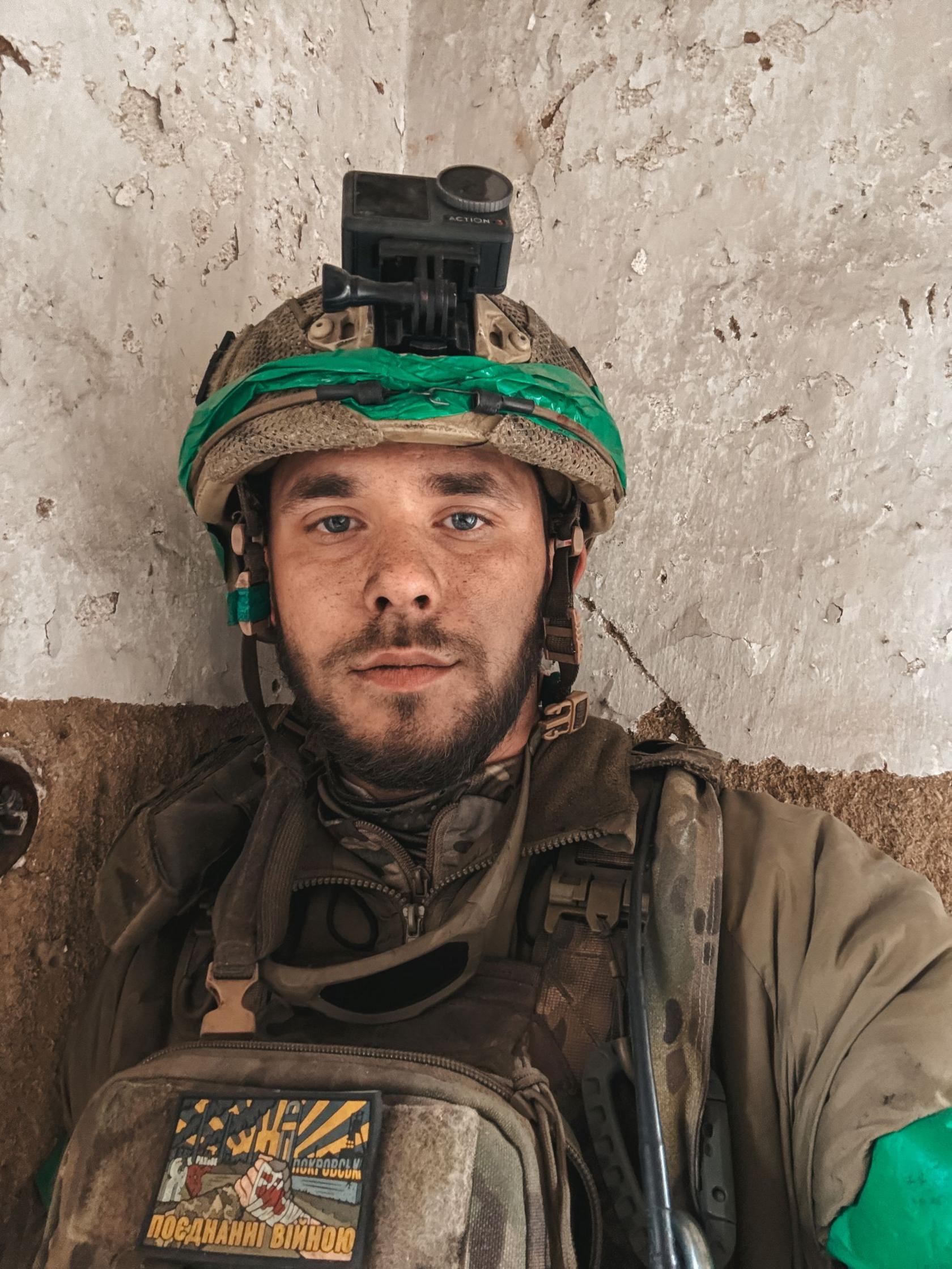 A selfie of a young man wearing Ukrainian military uniform. He is wearing a helmet with a camera on top. He has a short beard and moustache. He has taken this photo against a bare concrete wall.