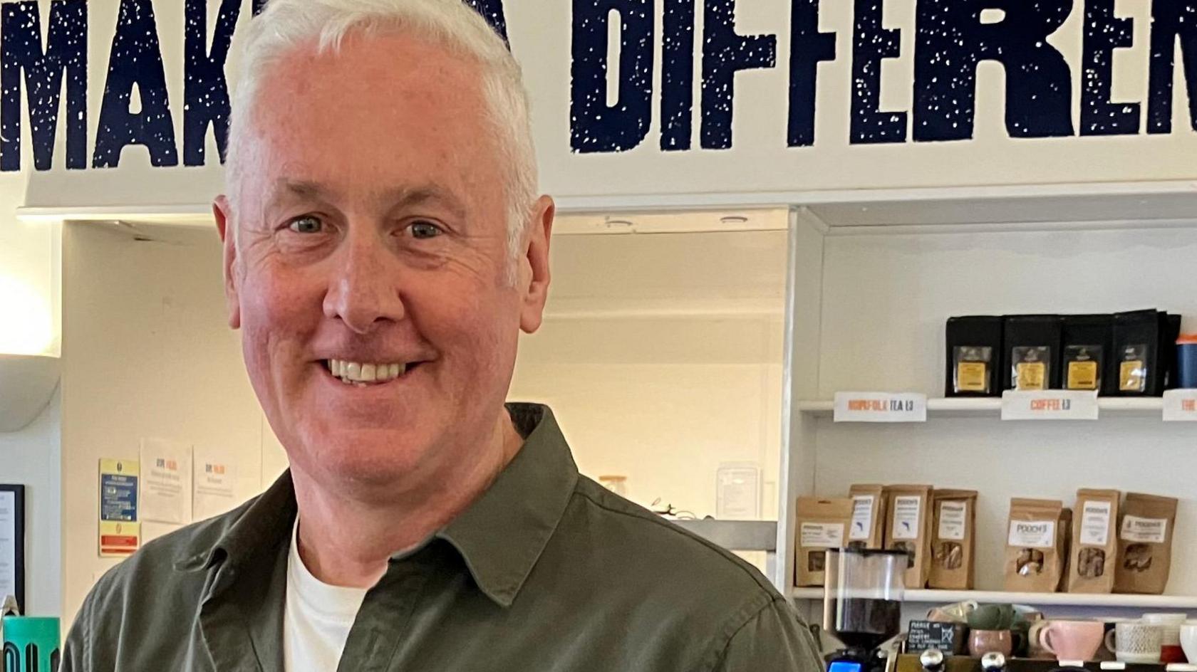 Chris Elliott stands in front of a coffee maker and stand containing slices of cake. He is wearing a khaki green shirt with a white t shirt visible under it. He has white hair. 