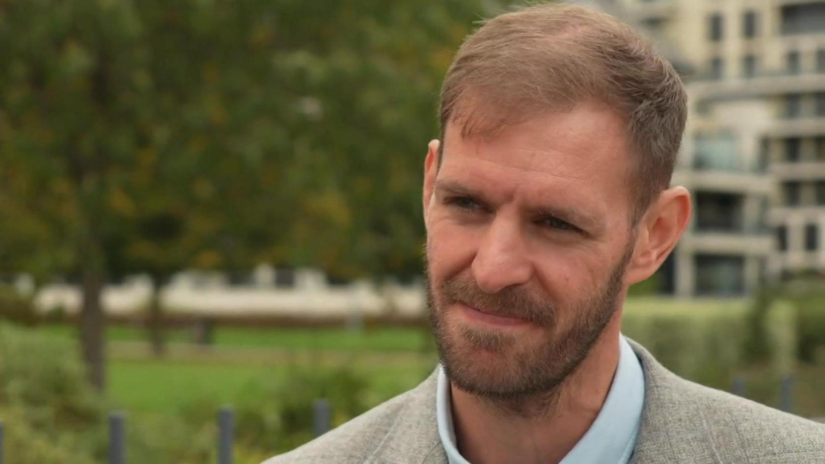Matt Hughes-Short in a grey suit with a blue shirt. He is standing in a park.