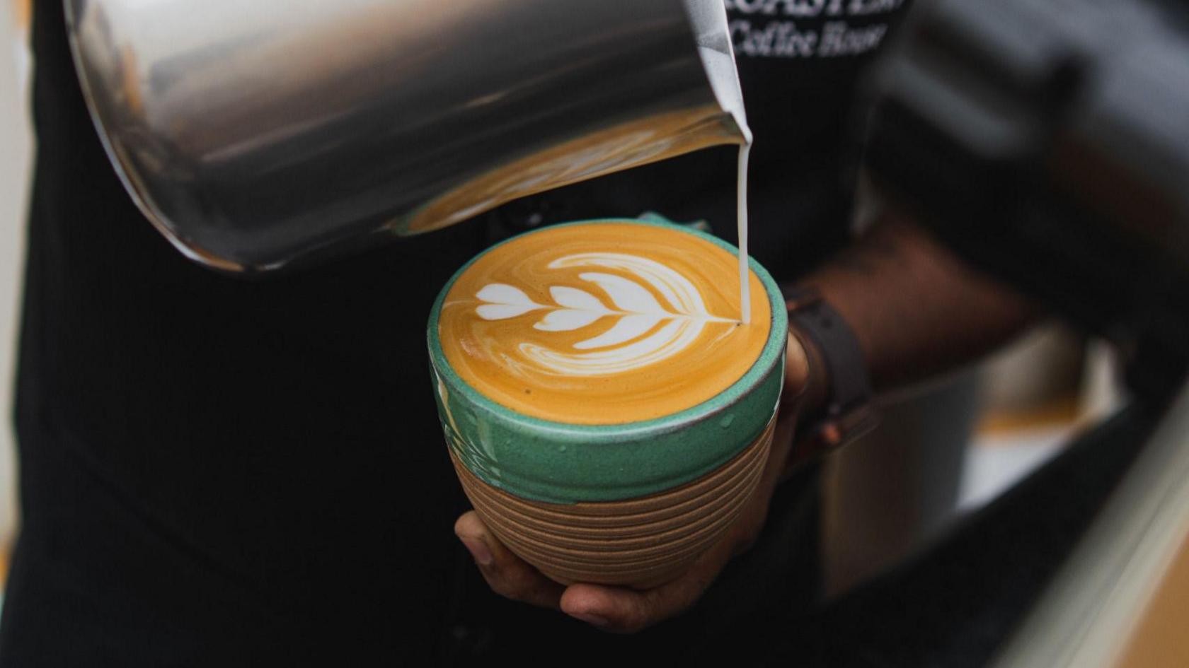 A barista crafting a milk foam design on top of a freshly brewed coffee in a mug