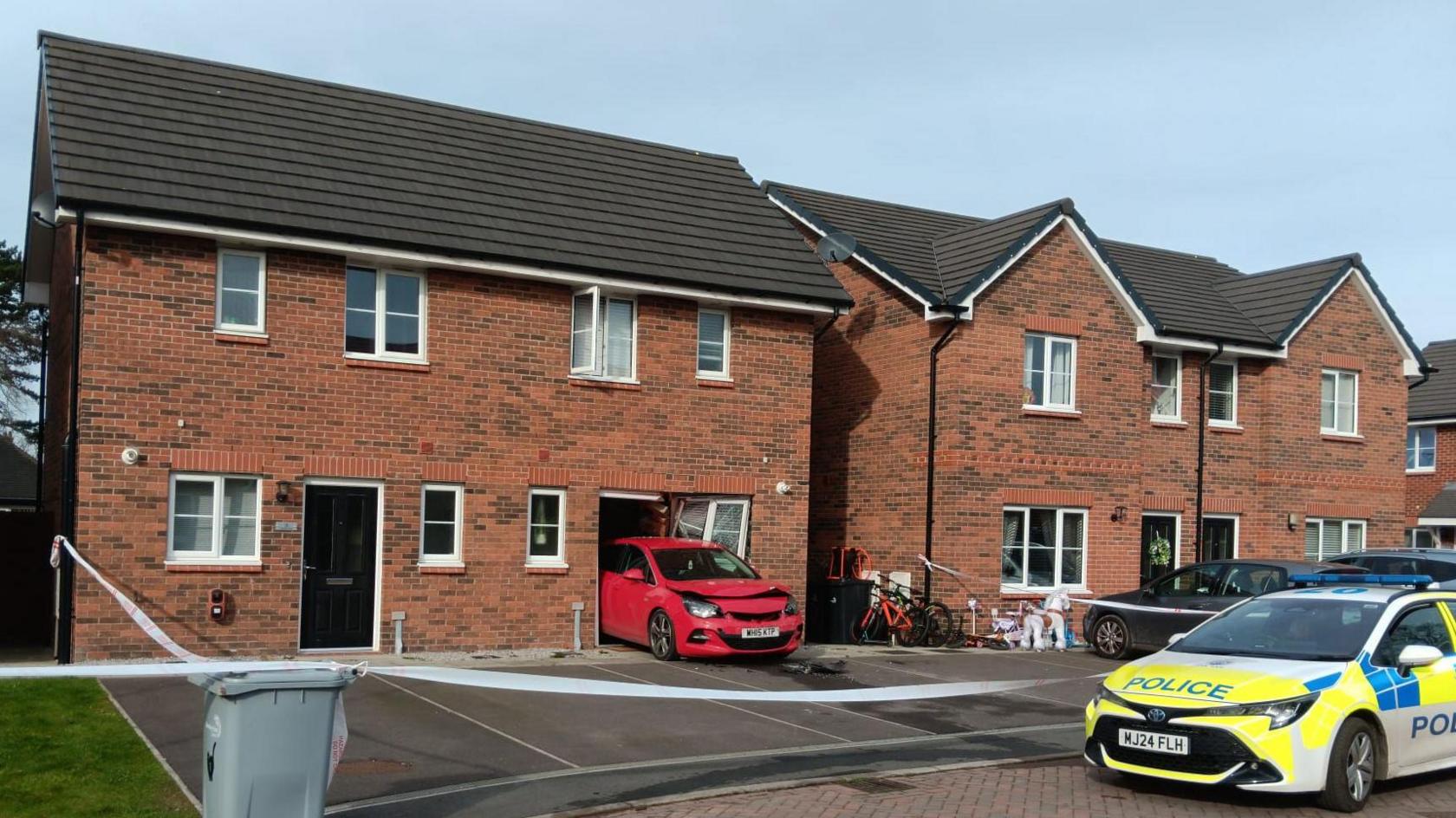 A row of semi-detached brick houses. A red car is sticking out of the front of one of the houses. A police car is sitting on the road outside the house.