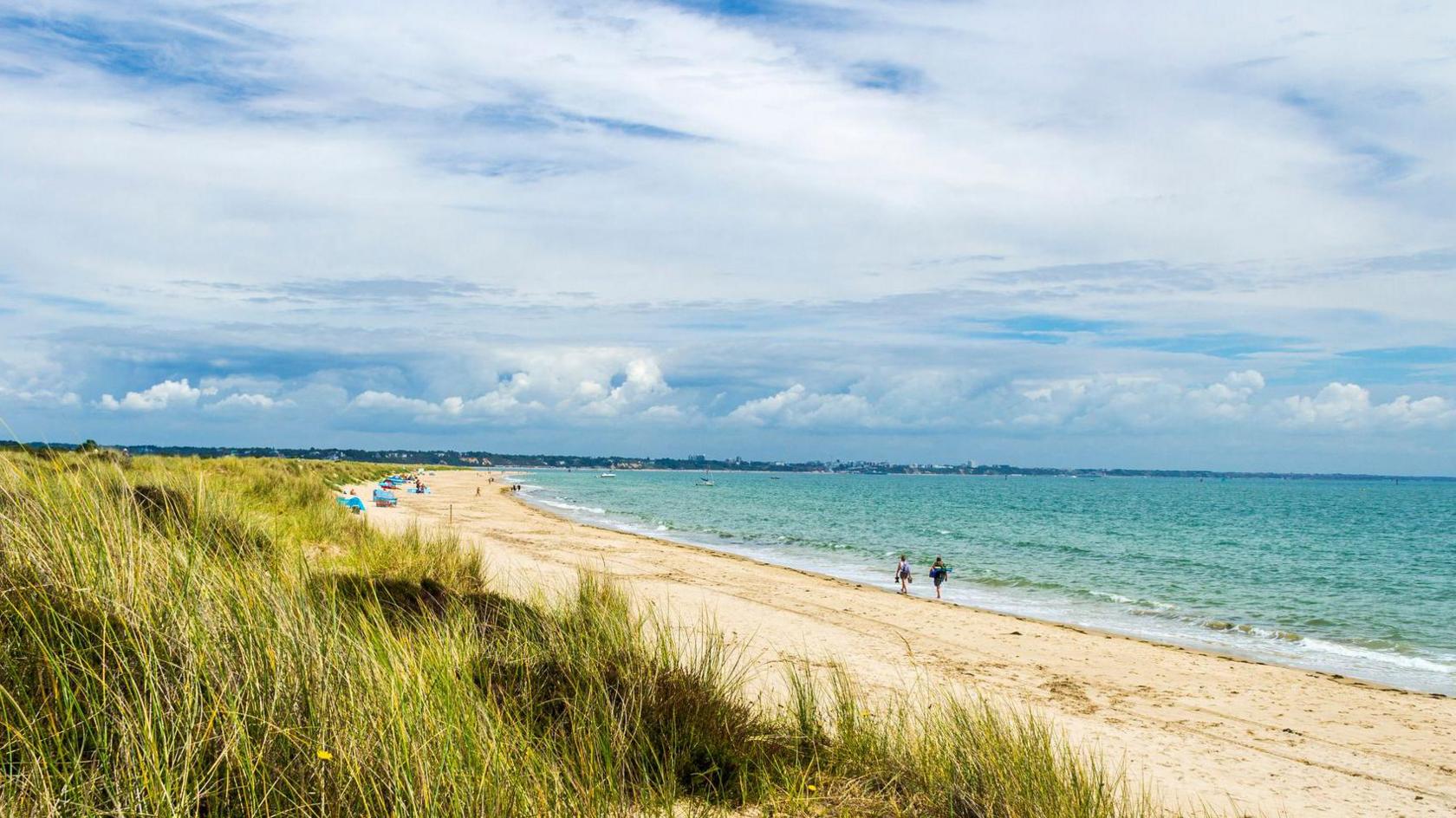 A sunny day at a beach. There is blue sky and golden sand with two people walking along the shoreline. You can see colourful windbreaks/tents in the distance and the beach has a strip of green dunes behind.