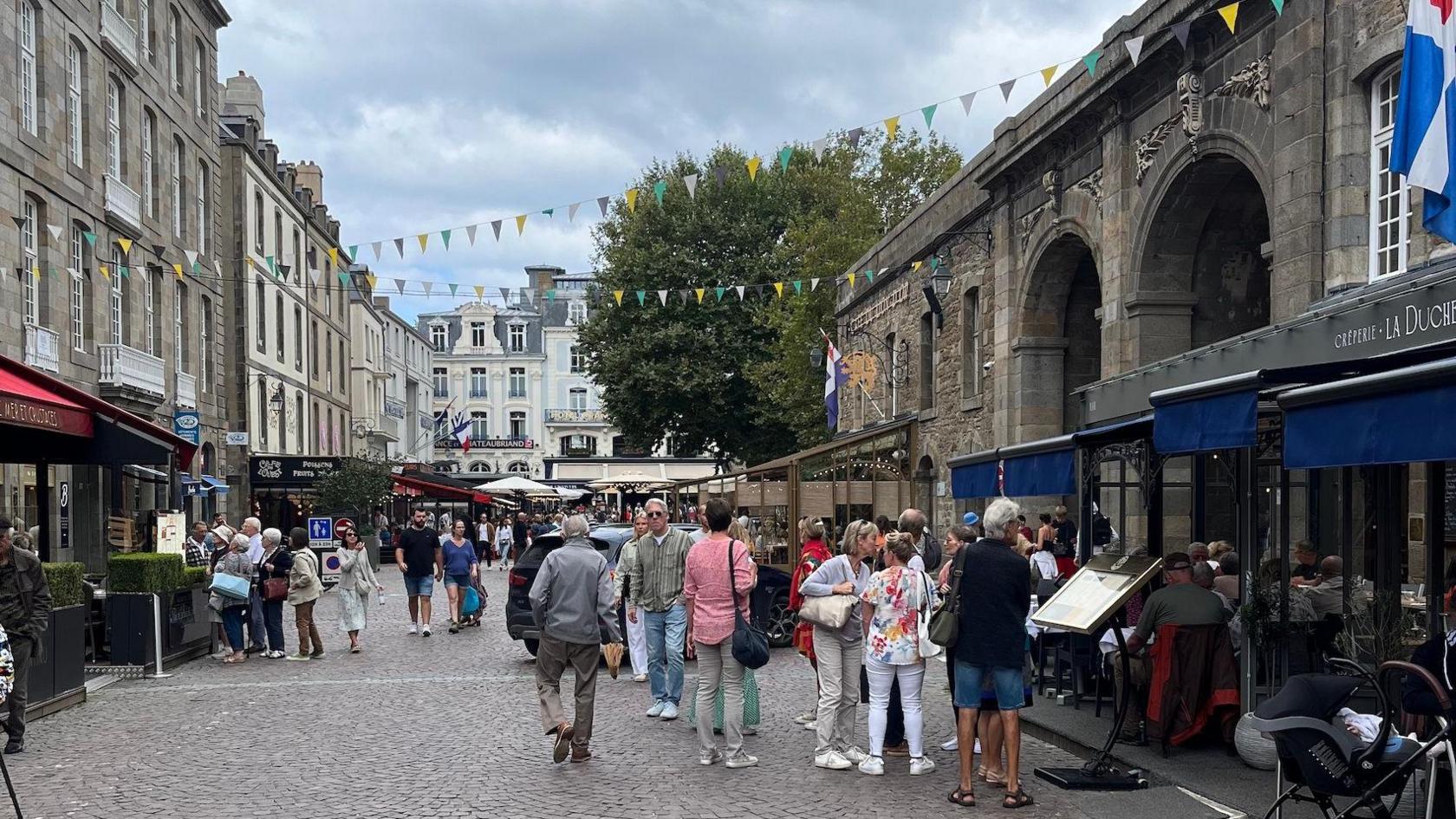 A crowded, cobbled shopping street in the historic French port city of St Malo, with granite-built cafes, restaurants and shops either side and bunting hanging above.