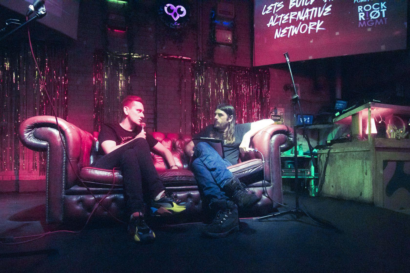 Andrew Miller, aka DJ Millabong, sits on a sofa with guest speaker Lyle Bignon. He has long hair and a beard. They are both in front of a screen which reads Let's build an alternative network.