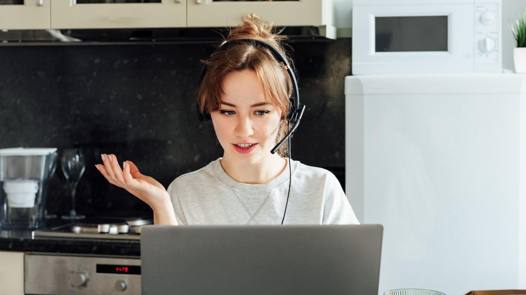 Woman sitting in her kitchen looking at a laptop, wearing a headset