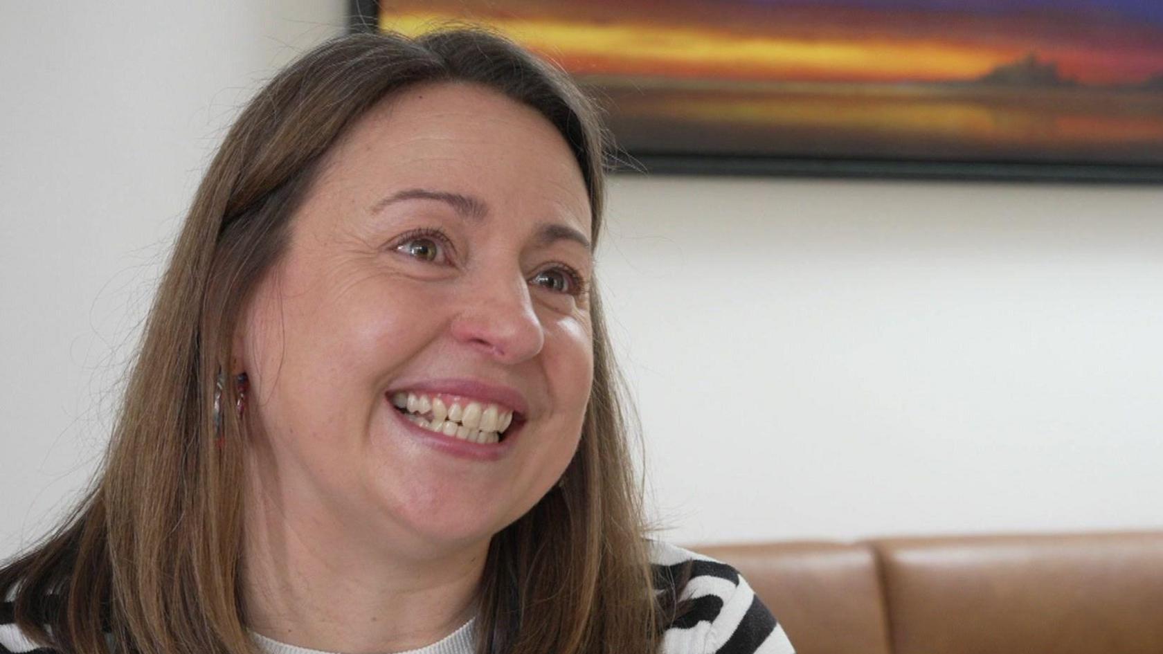 Georgina smiles as she talks to a reporter. She has straight, shoulder length brown hair and is wearing a black and white hooped top.