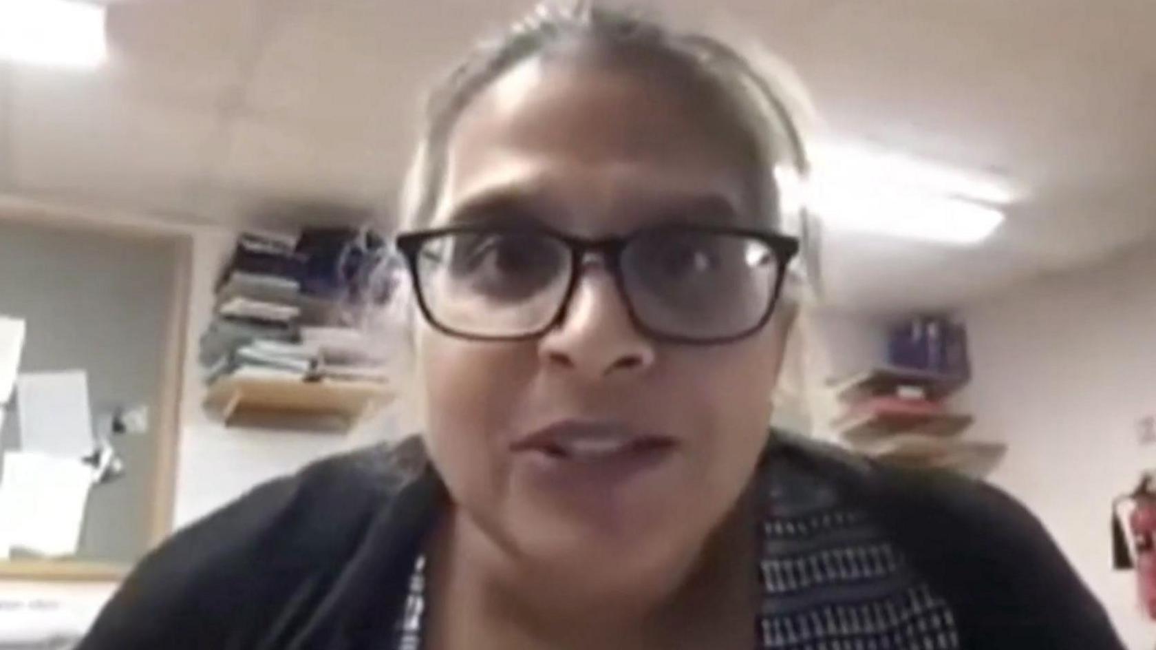 Kuldeep Stohr looks at the camera during an NHS video she is taking part in. She is wearing a black and white blouse and a black cardigan. She has black, plastic-framed glasses on and greying hair which is tied back. She is sitting in an office with folders packed onto shelves in the background and some papers pinned to a board.