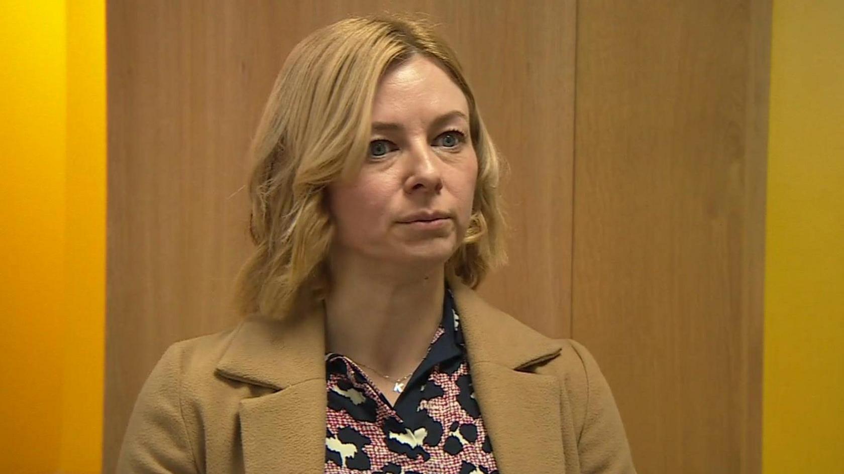 Kenna Young looks slightly off camera, she stands infront of a wooden door and yellow painted walls. She has shoulder length wavy blonde hair and wears a beige wool collared coat and a black, red and white leopard printed blouse