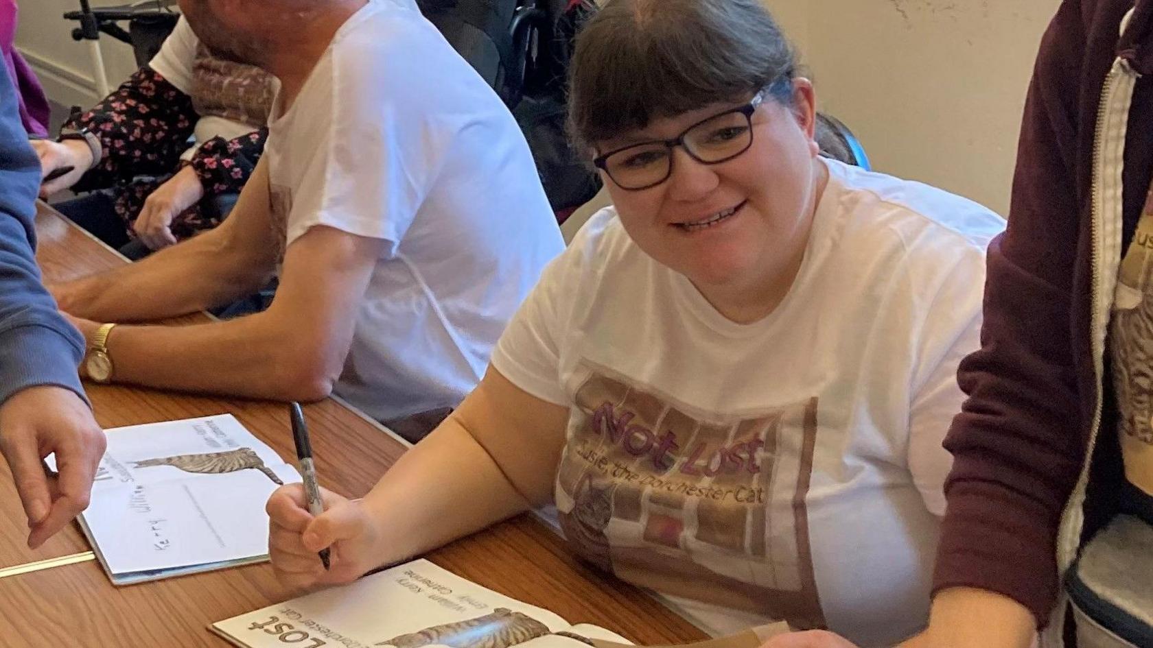 A smiling Emily Burr signing copies of the book at the launch. She has dark hair with a fringe and a ponytail and is wearing dark-rimmed glasses.