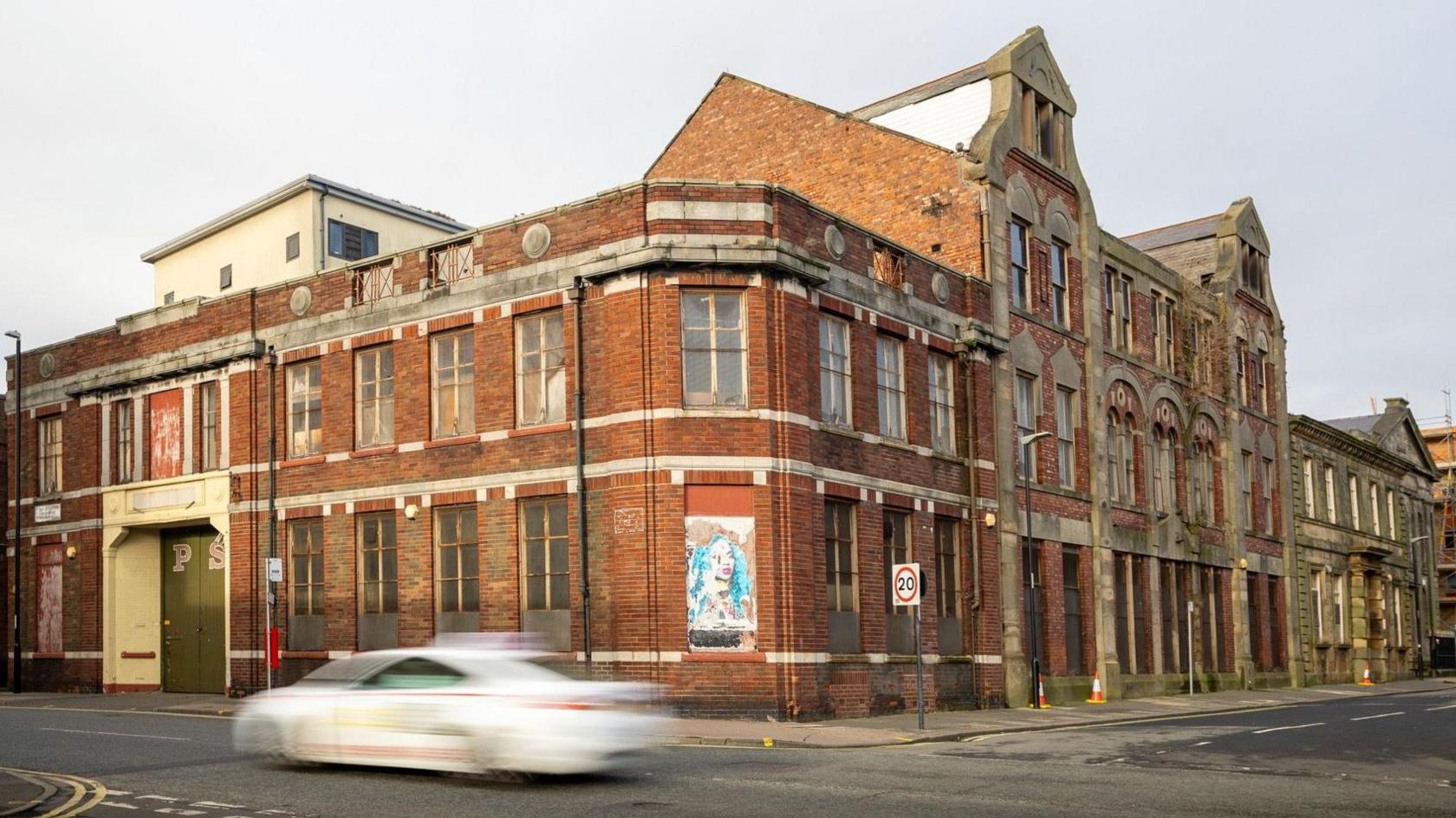 The former Peter Smith Antiques building. It is constructed with red bricks and is located on the corner of two streets. Some of its windows are boarded up. A white car is being driven past.