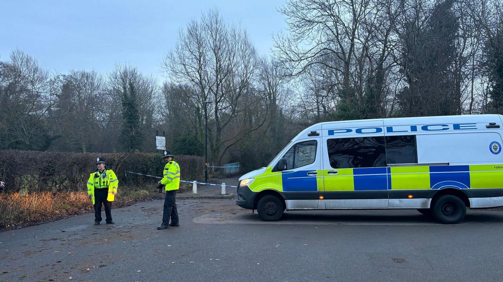 A blue and white police cordon is stretching across a path. There is a white, blue and yellow police van parked in front of it. Two police officers are standing near the van in yellow police jackets and black police hats.