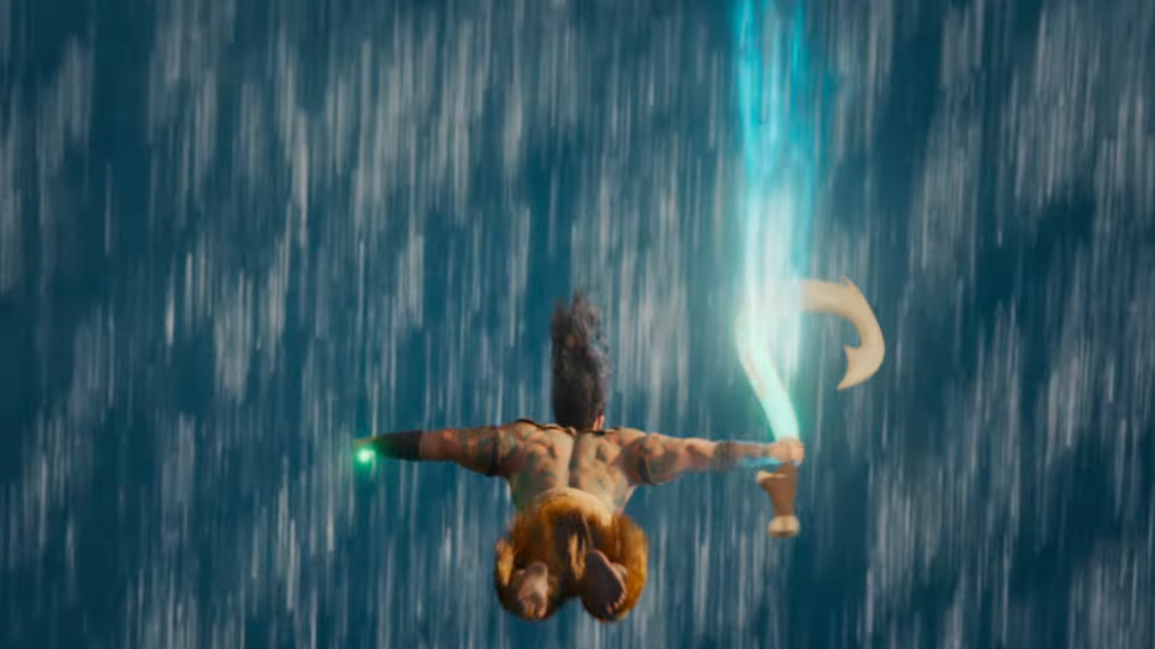 Screengrab from the trailer showing Maui facing from the camera as he falls in front of a waterfall. In his right hand his has a large fish hook and in his left hand his had a glowing green object. 