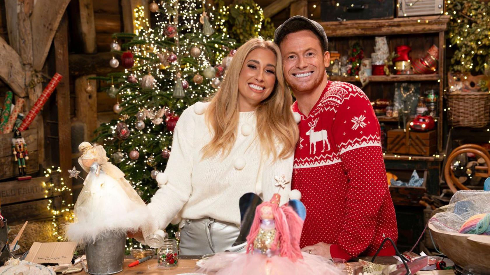 Stacey and Joe pictured on the set of Stacey Solomon's Crafty Christmas, in a farmhouse type room with a large Christmas tree behind them and shelves piled with boxes, baskets and fairy lights. Joe wears a red Christmas jumper and grey cap, while Stacey wears a white fluffy jumper. 