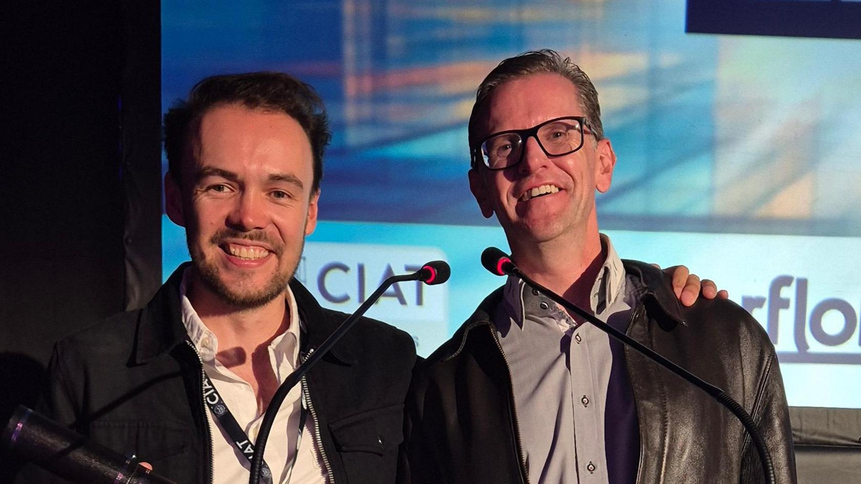 Two men wearing black jackets and smiling in front of a podium, holding a black award