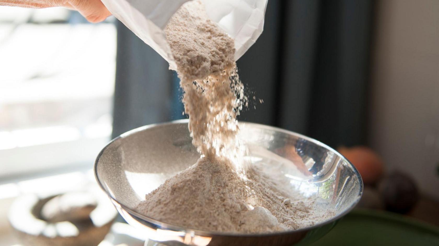 A person pours flour from a white bag into a silver baking bowl.