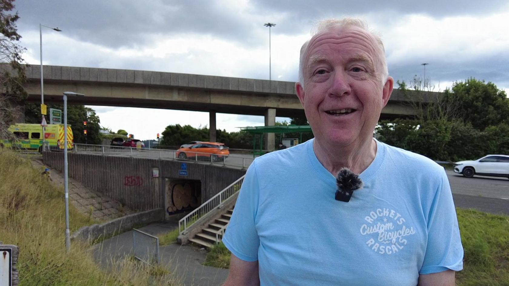 Mark Swarbrick, an older man wearing a light blue T-shirt, stands in front of an A Road roundabout in Plymouth, which also features an overpass and an underpass.