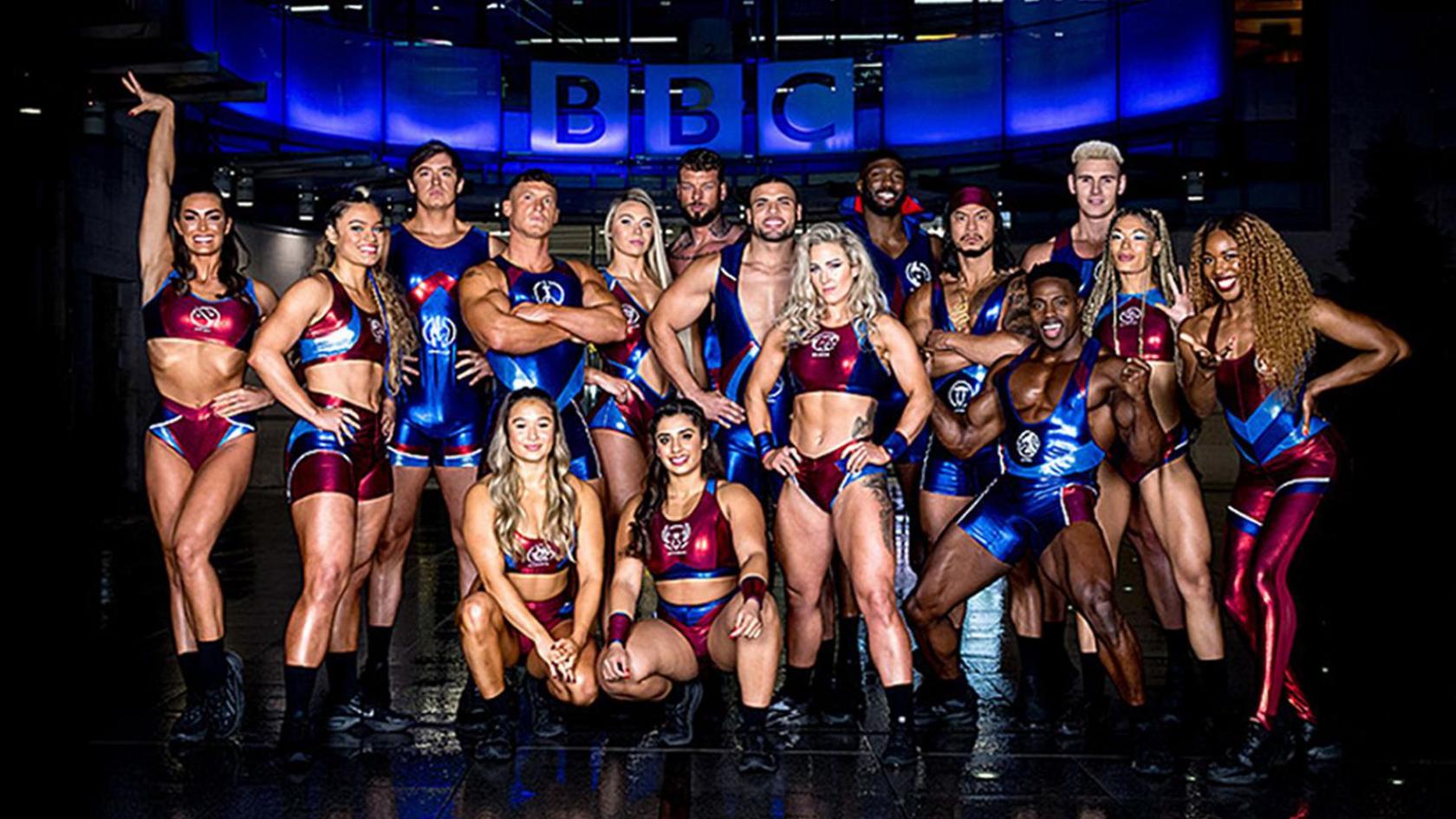 A promo shot of 16 Gladiators in their shiny red and blue lycra costumes posing beneath a blue illuminated BBC logo.