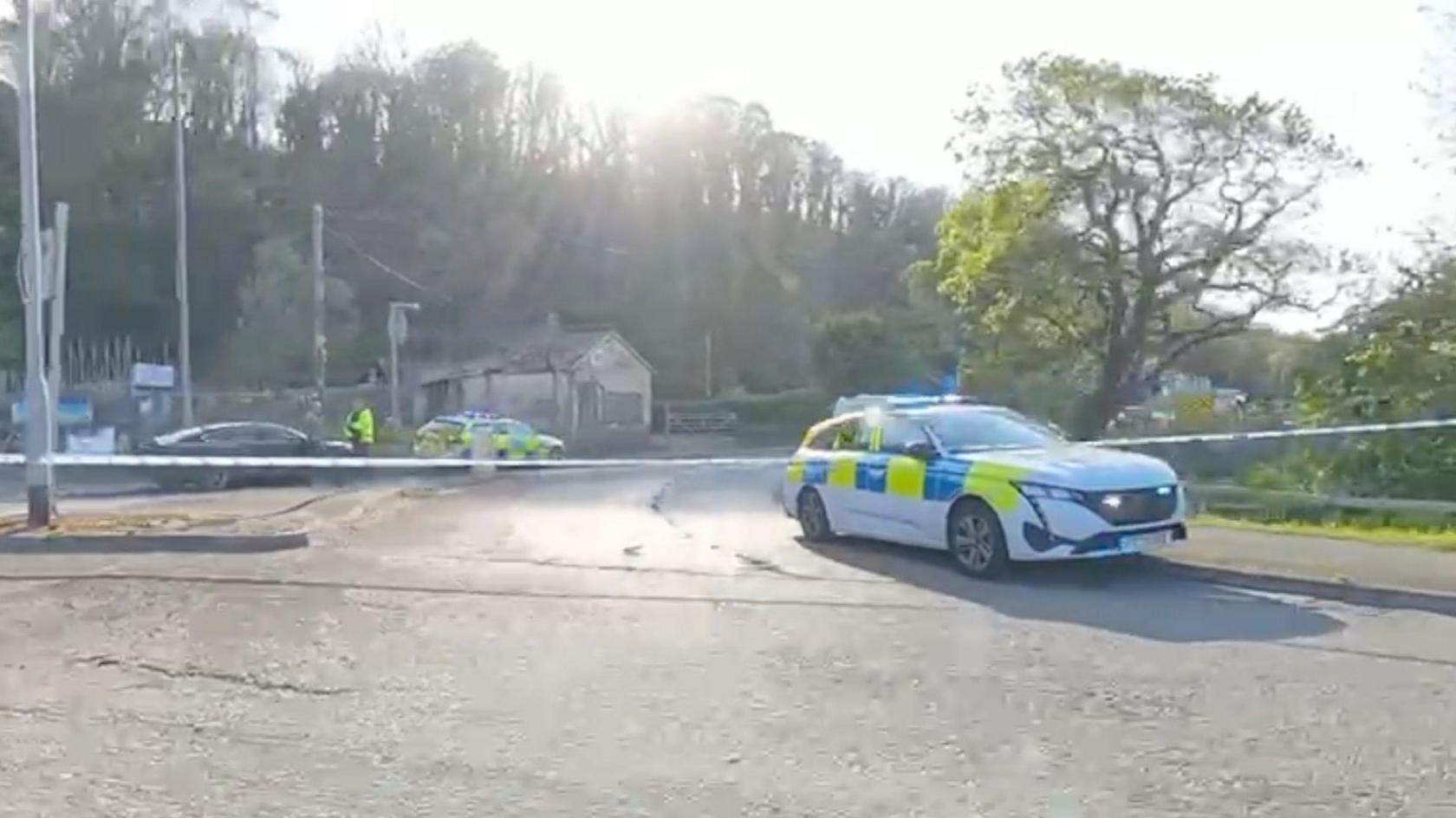 The location of the crash in Flintshire. The area has been cordoned off by police tape and a police car. Another police car can be seen in the background with one police officer talking to a man by a black car.
