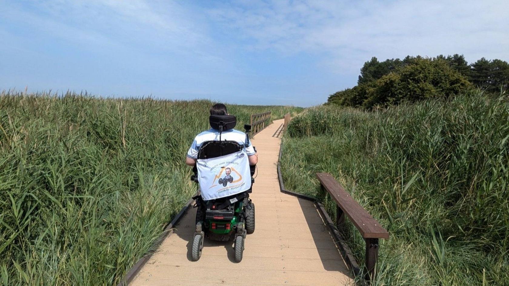 A man in a wheelchair is shown moving away from the camera using a wooden boardwalk that has long reeds on either side.