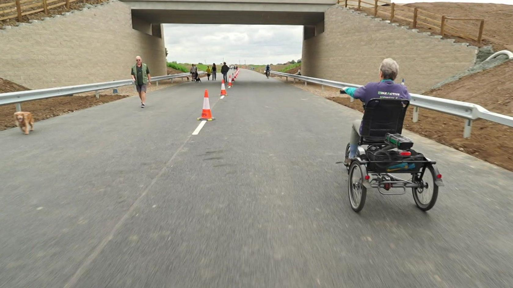 A woman on a bike rides towards a bridge crossing the new road, while a man walking a dog walks on the other side of the road.