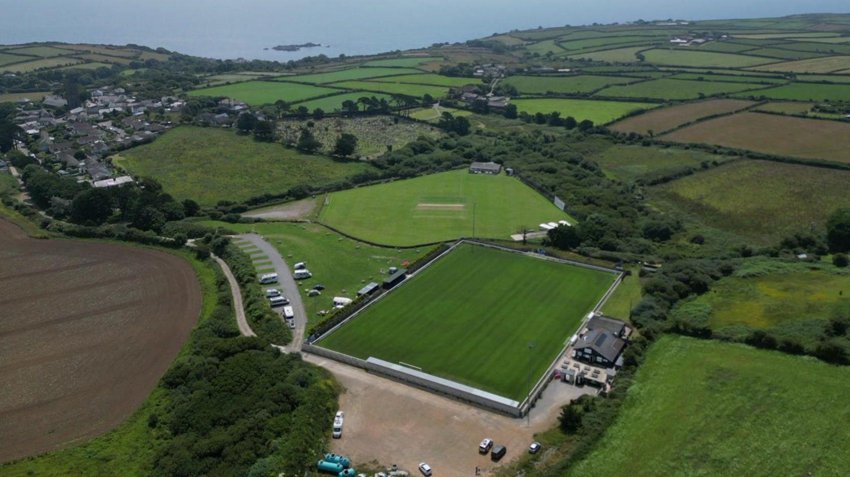 A picture taken by a drone of a green, rural landscape with lots of fields and a small village in the distance. The sea is also in the distance. There is a football pitch and cricket pitch at the centre of the picture.