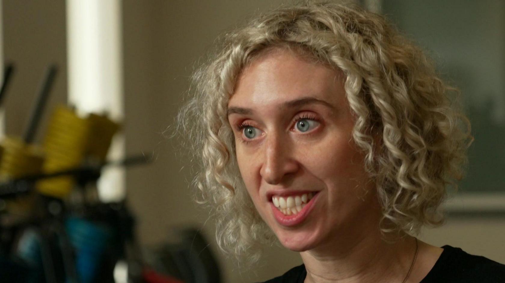 A woman with curly blond hair sits speaking to someone off camera.