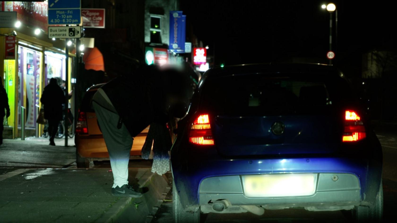 A blue car pulled up to the curb on a street at night. A woman is talking to the people in the car. Her face is blurred and it is dark, so she is hard to make out.