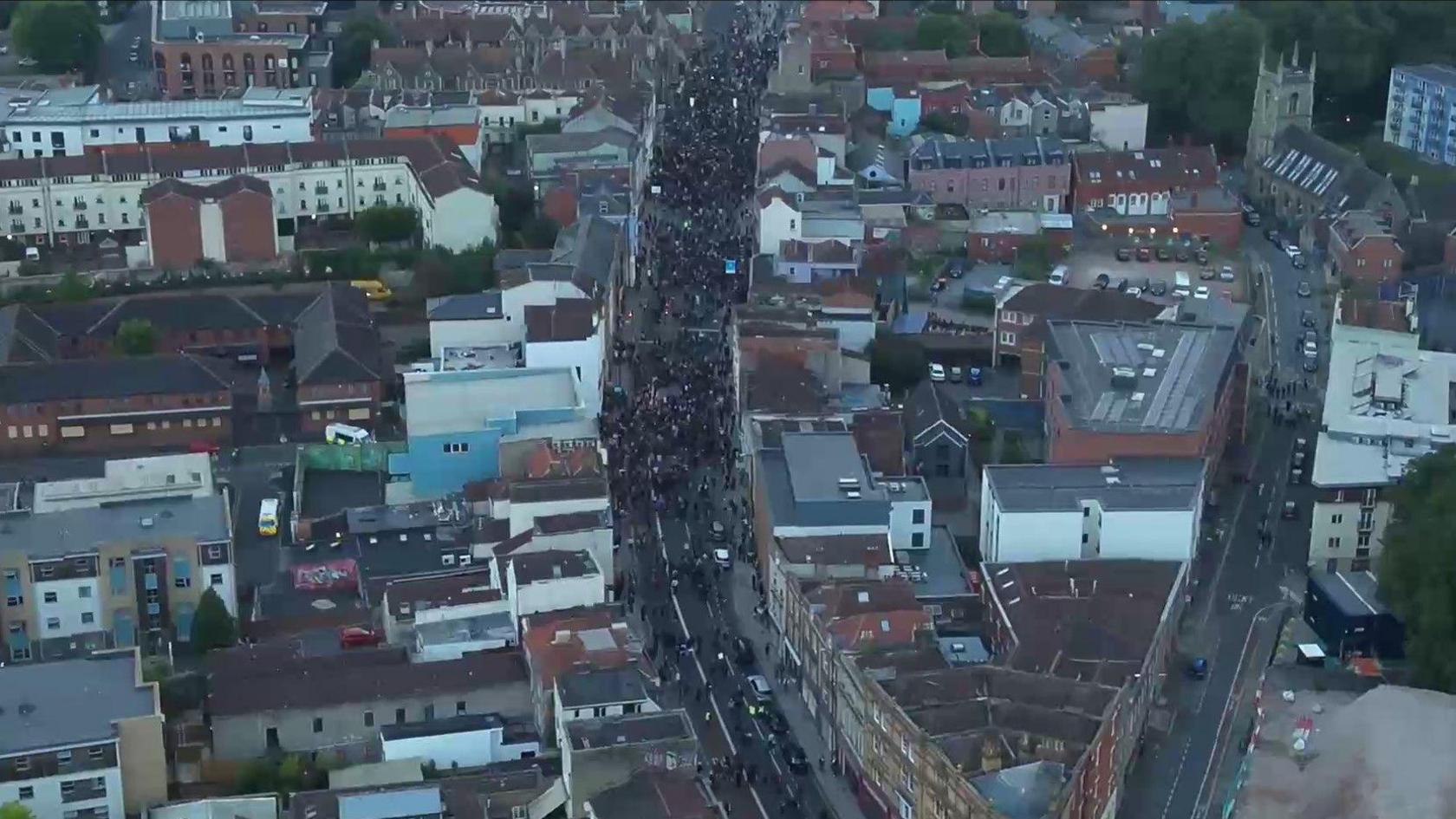 An aerial shot of Old Market taken from a helicopter at dusk. City buildings can be seen on either side of a main street - West Street - on which hundreds of people can be seen gathered. 