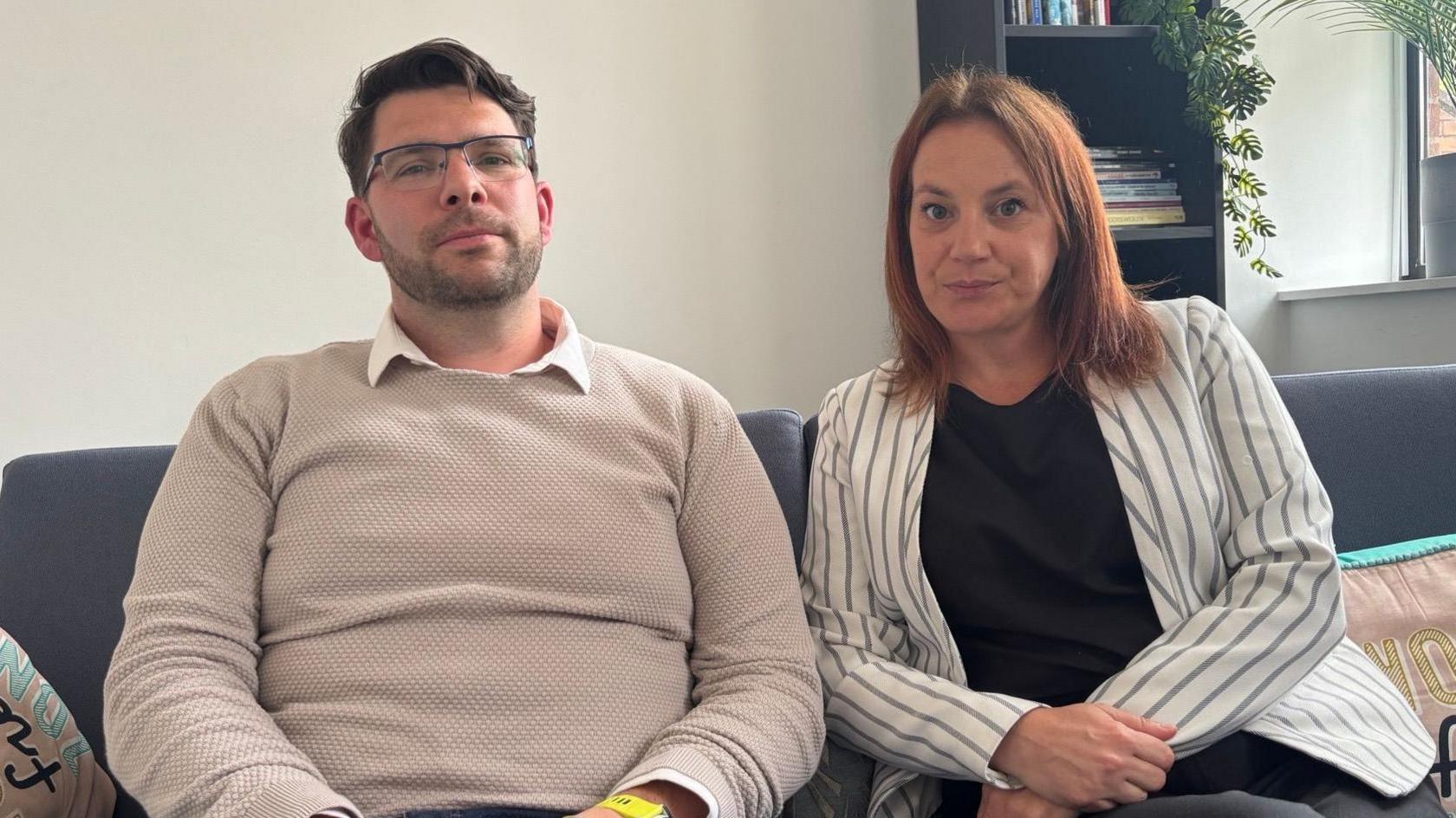 The two parents are looking straight into the camera. Michael has a cream jumper on whilst Kirsty has a striped suit jacket on an black top. They are sitting on a sofa with a book case in the background and some plants.