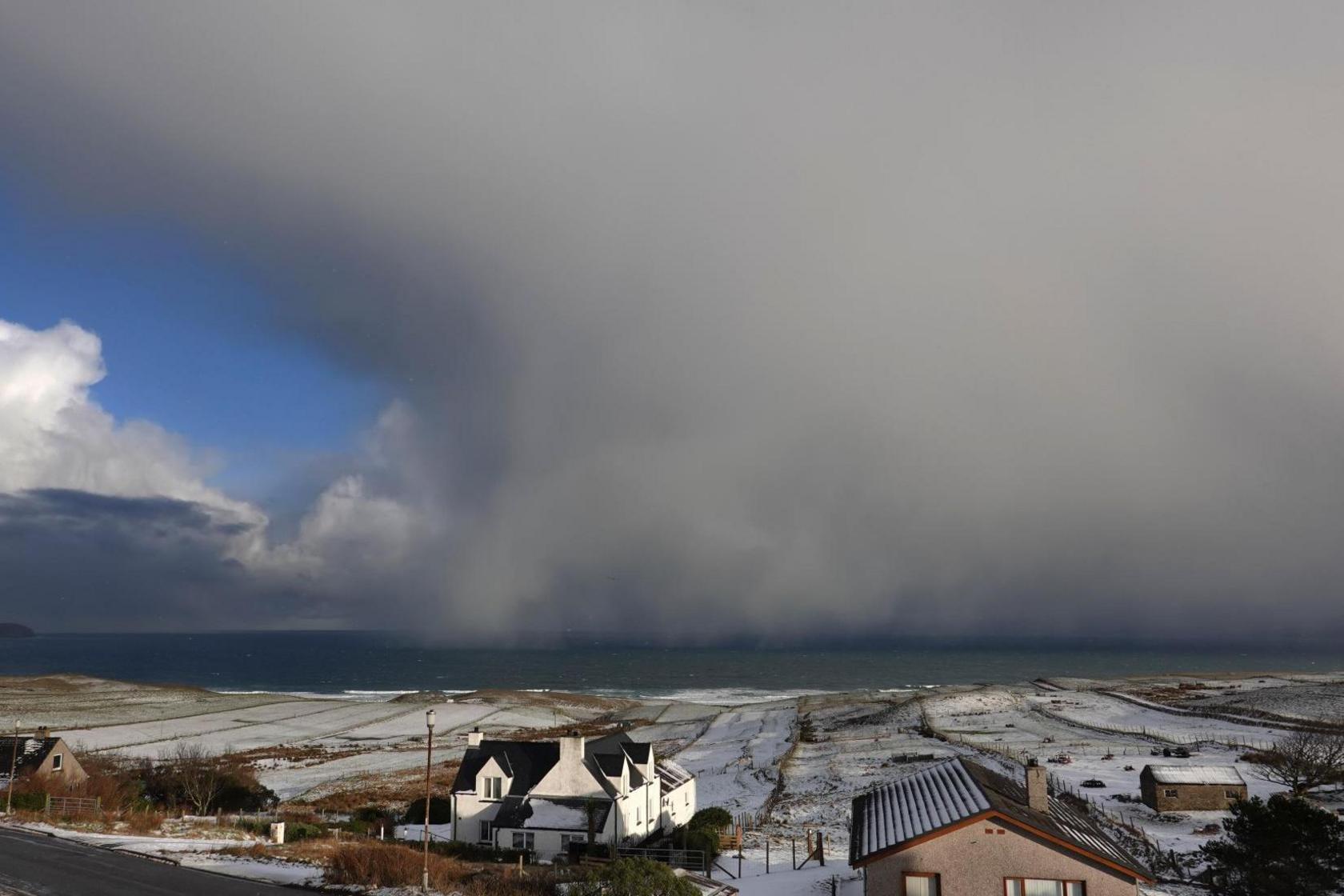 Three houses and a shed on an island coast. The sea is a grey-green colour below a shower of heavy snow.