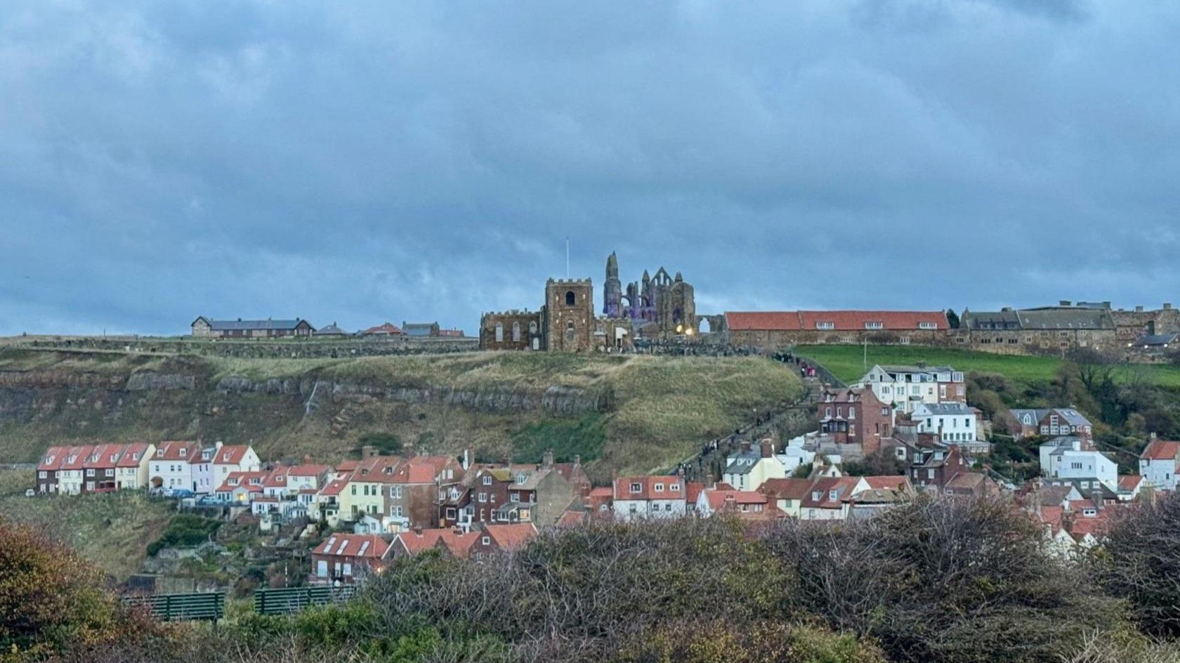 A ruined abbey in the distance behind a church. Low rise buildings near the abbey, which is on a grassy clifftop. Buildings in the valley beneath the clifftop.