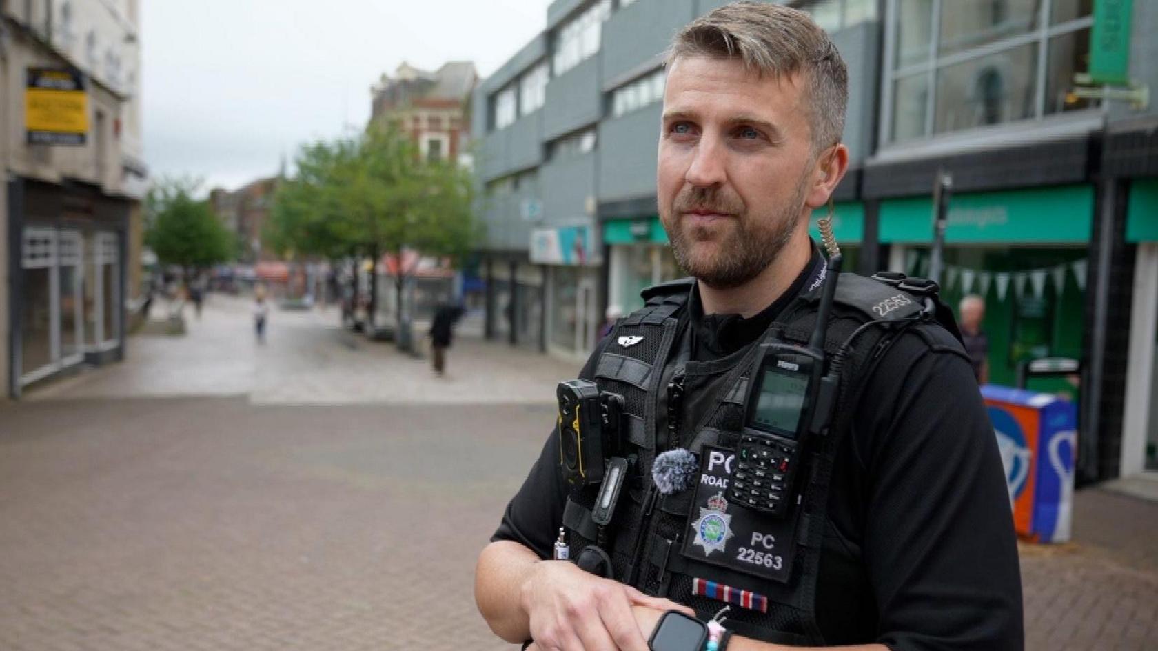 PC Tom Cordell stands in a street. He is a police officer in uniform, with short brown hair, parted to the left, and a short beard.