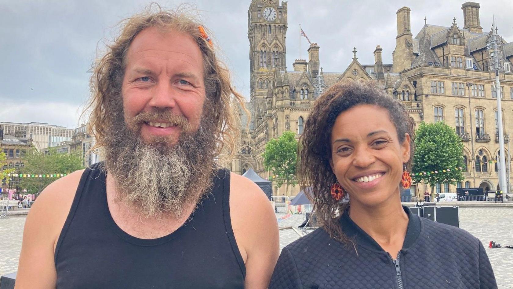 A man with a beard wearing a black vest standing next to a woman with dark brown hair and wearing a black, zip-up top with Bradford City Hall in the background.