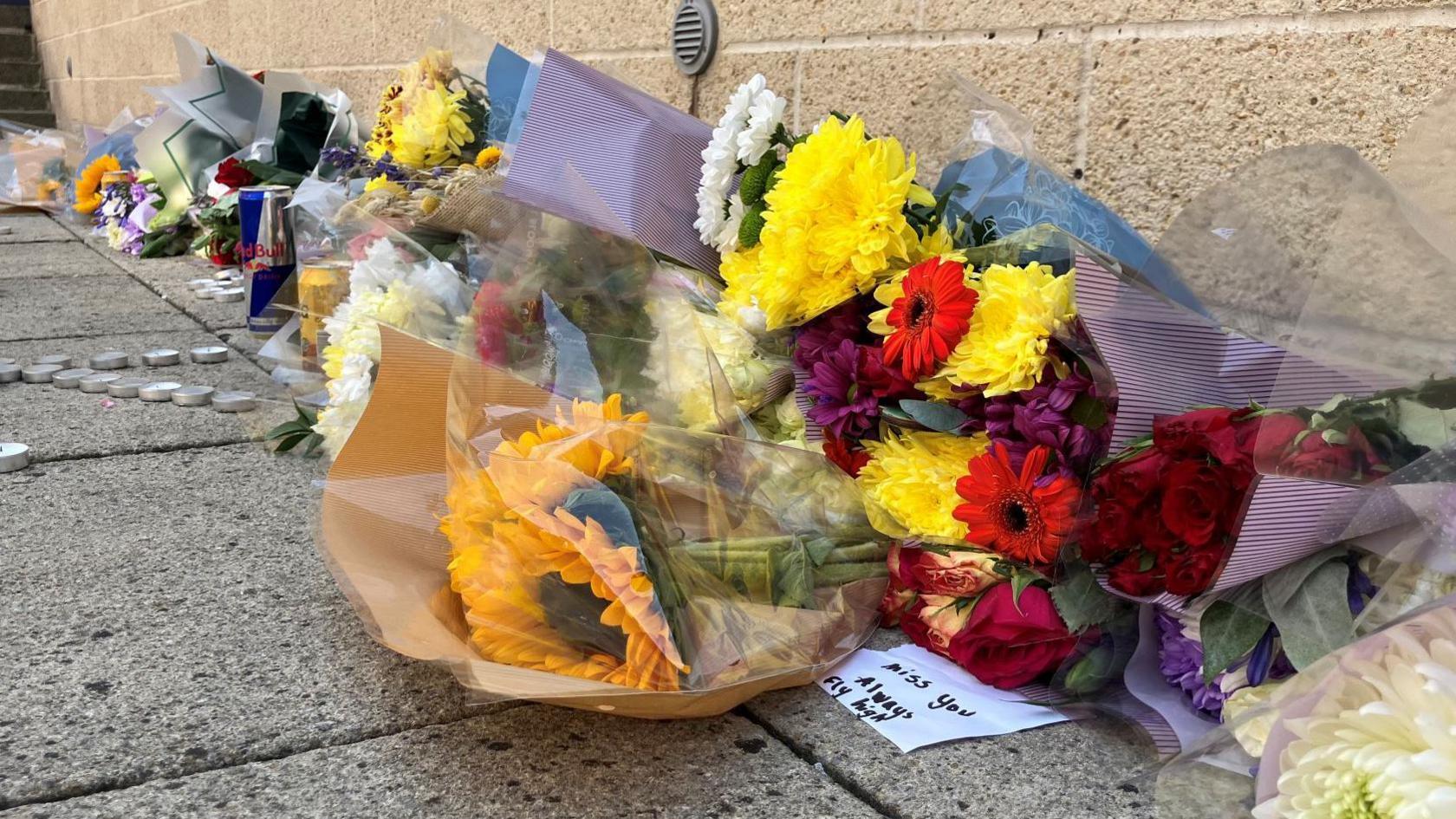 Bunches of flowers, candles and tealights are placed against a wall. Cans of drink are left too. 
