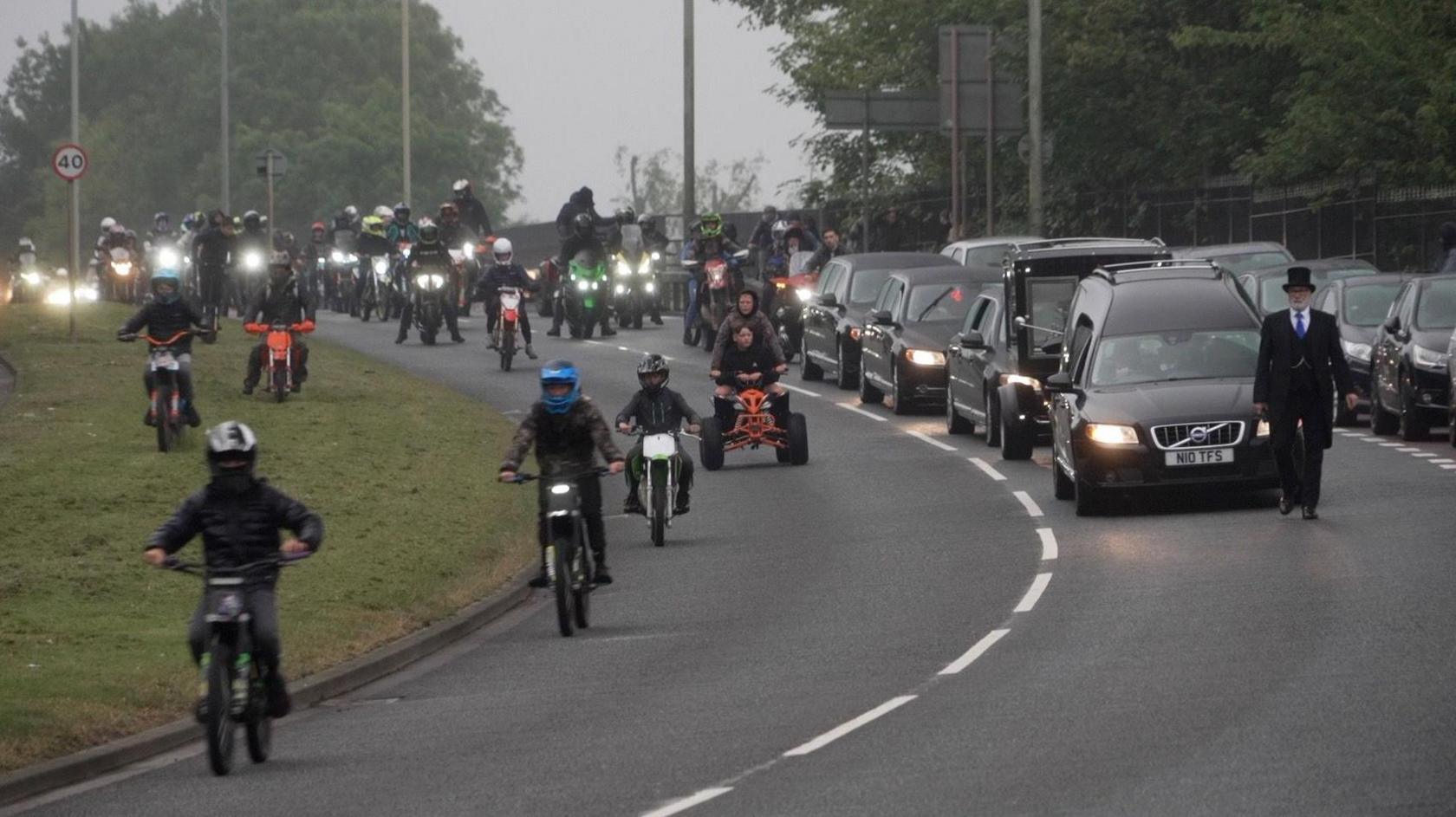 Dozens of bikers and youngsters on scrambler bikes ride alongside the funeral cortege. The bikes and cars have their lights on in the gloom.