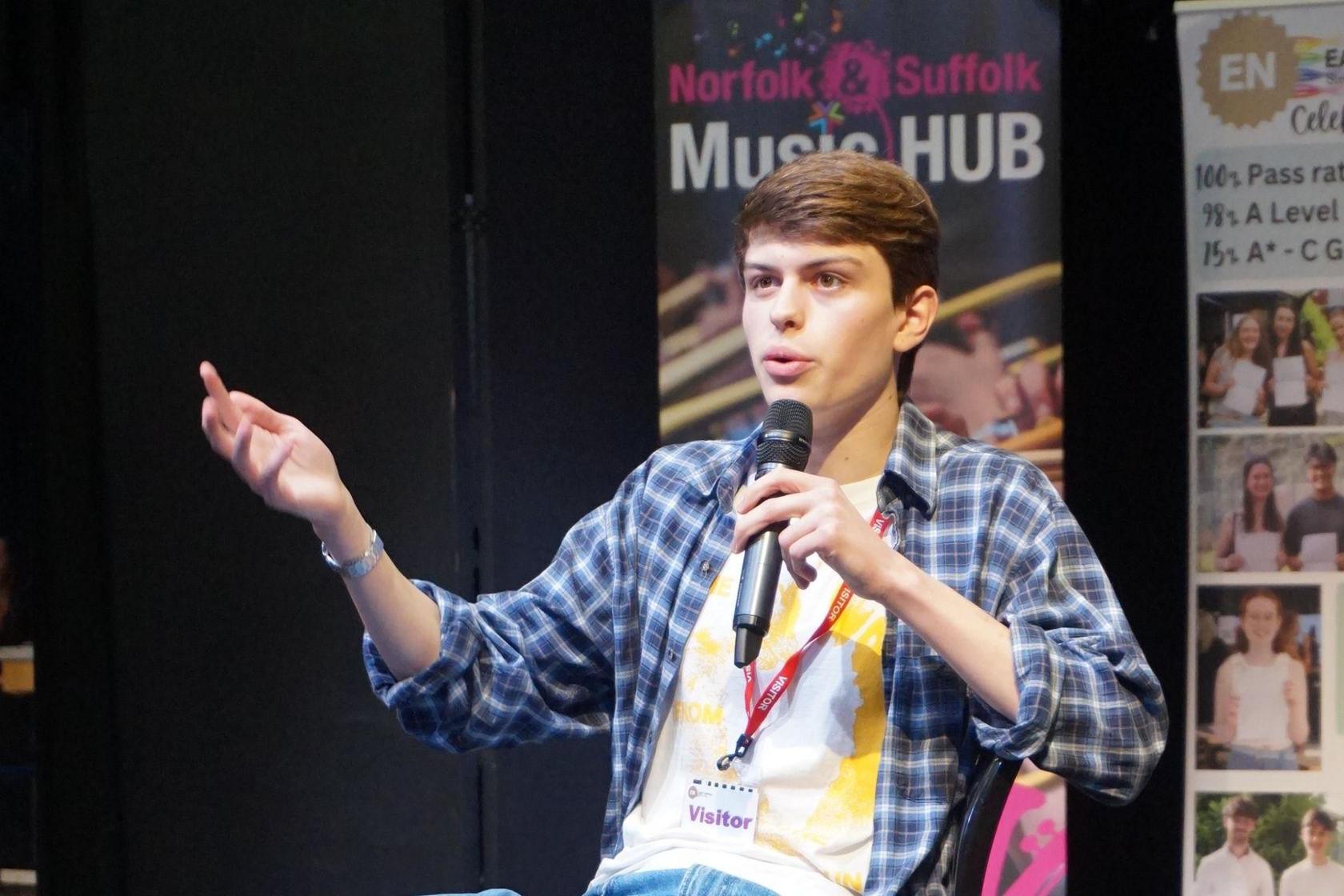 Tom Major sitting on a chair on a stage with a microphone in his left hand.