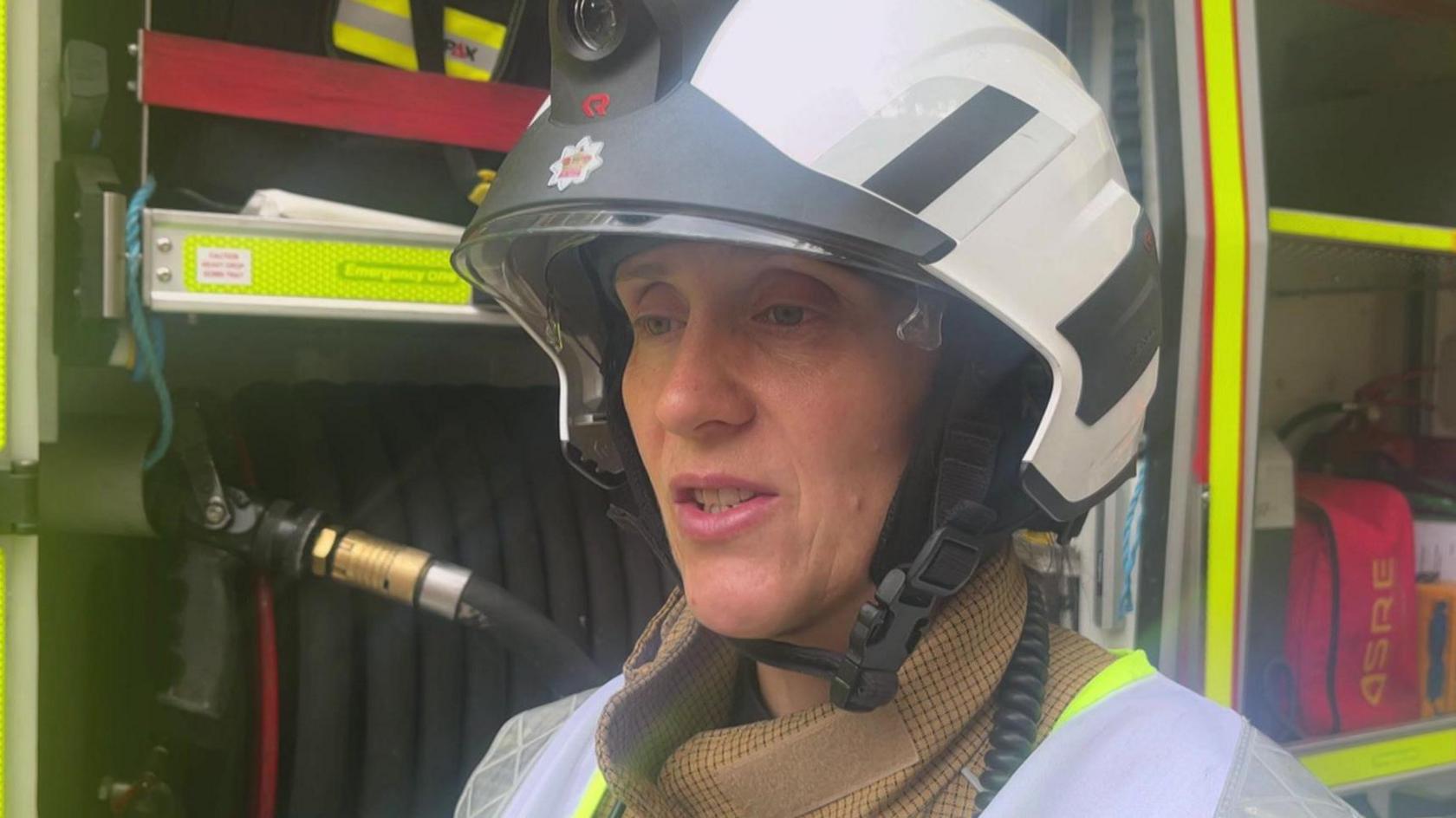 A woman firefighter wearing a white and black helmet, brown uniform and hi visibility jacket. She is standing next to a fire engine.