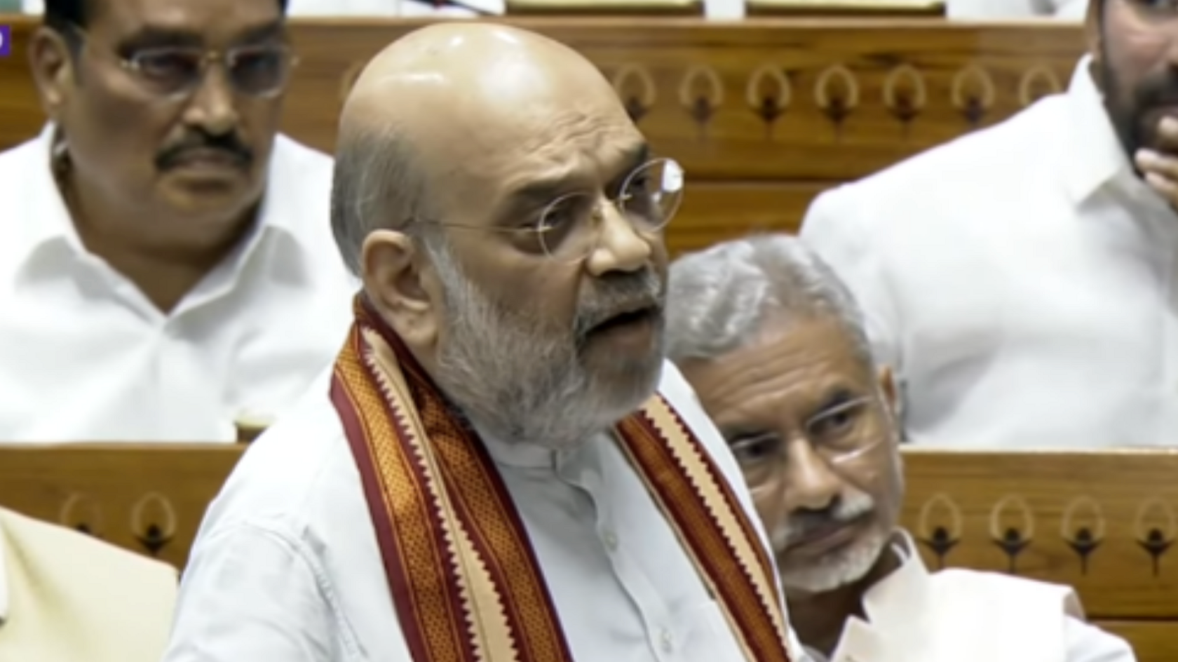 Amit Shah seen in a white kurta with red and gold scarf around on his neck as he speaks. Members of the ruling BJP seen sitting in benches behind him.