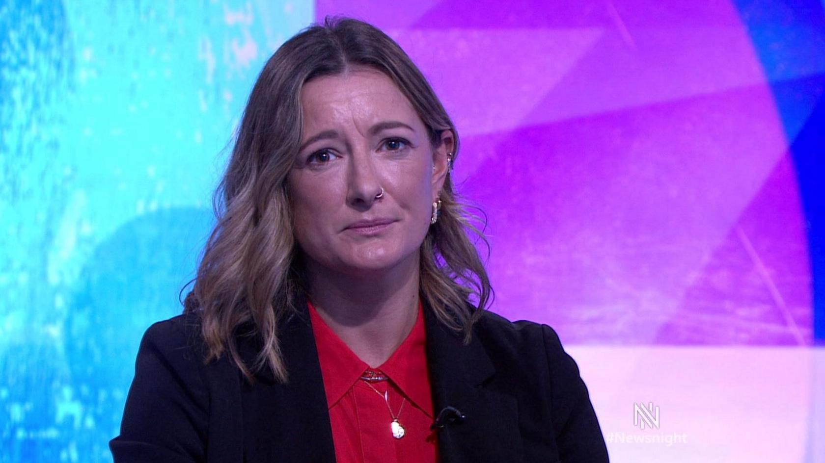 Image shows Morwena sitting in front of the purple and blue newsnight background wearing a red shirt, black blazer.